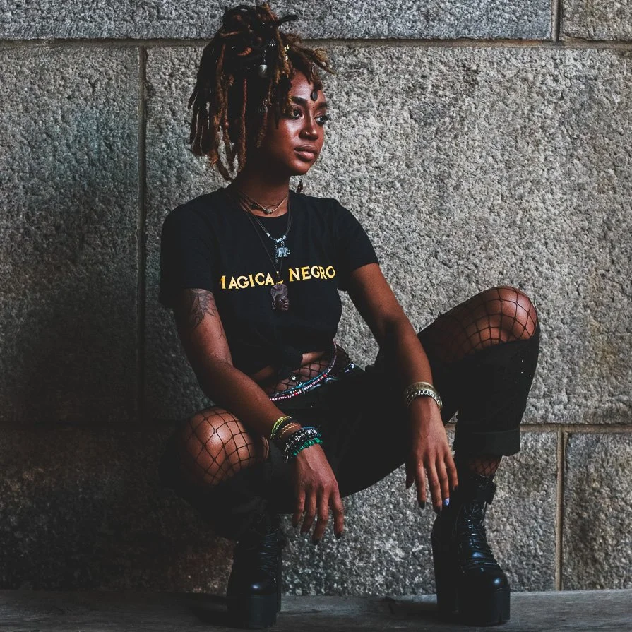 A young woman with dreadlocks sitting on the ground against a rough stone wall, wearing a black T-shirt, fishnet stockings, black boots, and accessorized with multiple necklaces and bracelets.