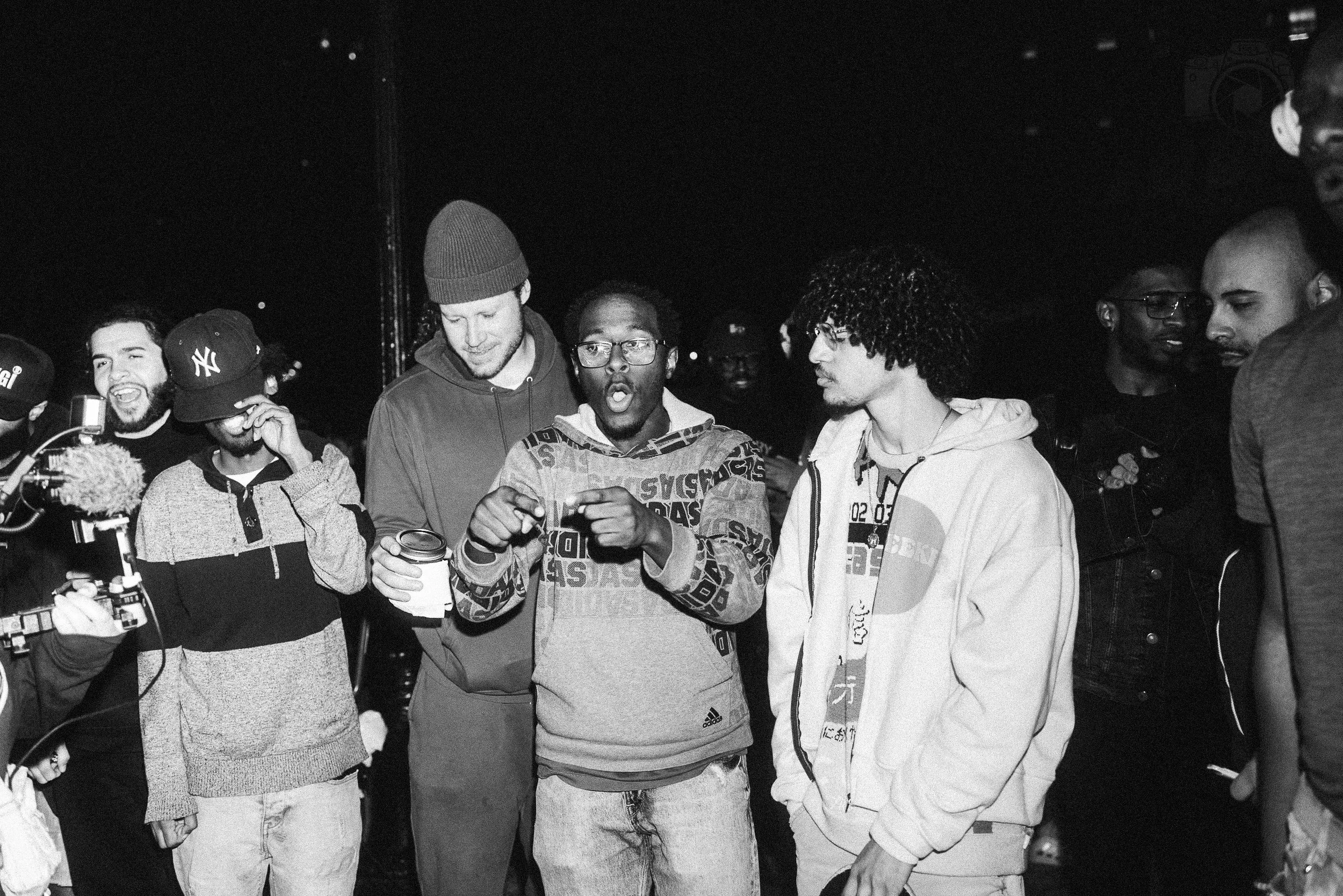 A group of young men gathered together outdoors during the night, engaged in conversation. Some are wearing hoodies and caps, and one is holding a drink. The atmosphere appears lively and social.
