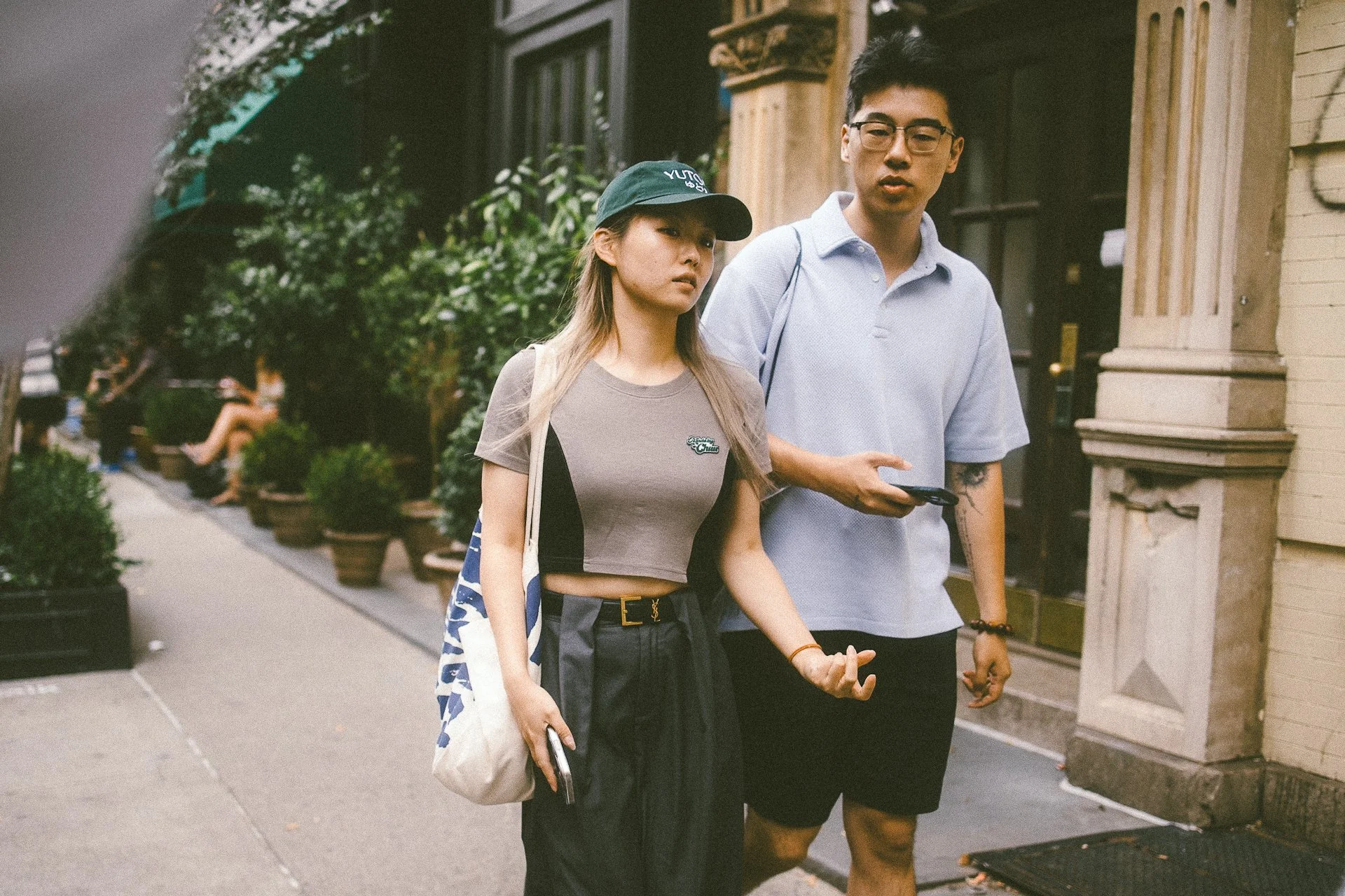 A young woman and man walking together on a city sidewalk, holding hands, with buildings in the background.