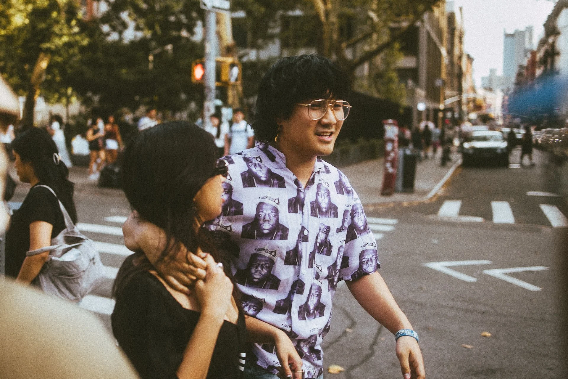 A man with black curly hair and glasses wearing a purple shirt with black and white faces printed on it, walking on a city street. A woman with dark hair and a gray backpack is partially visible in the foreground. There are several people and cars in