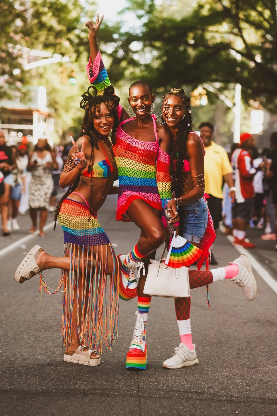 Three women in rainbow-colored outfits celebrating at an outdoor pride event, with a crowd in the background.