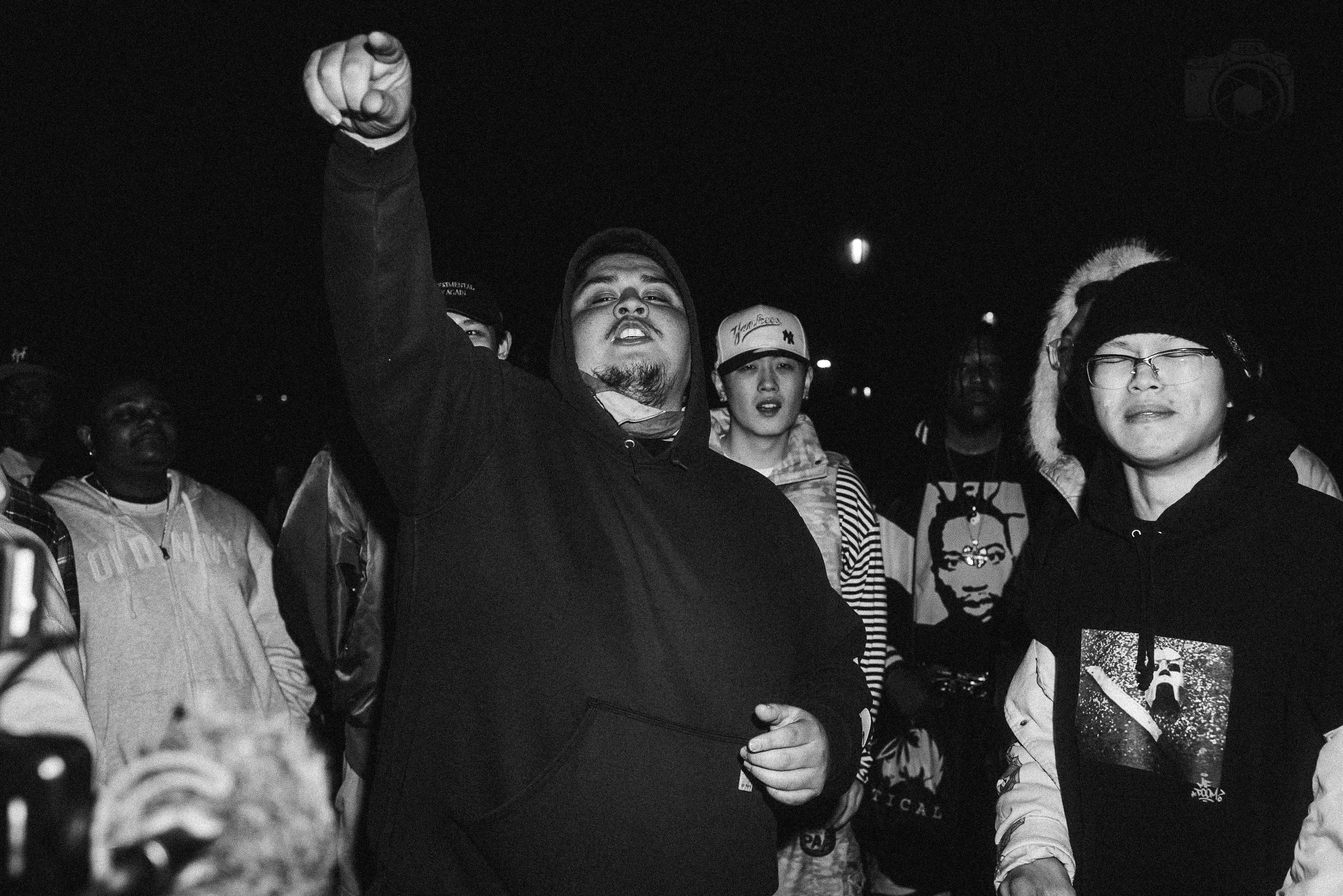 Group of young protesters outdoors at night, some wearing hoodies, jackets, and hats, with one person pointing and others holding signs.