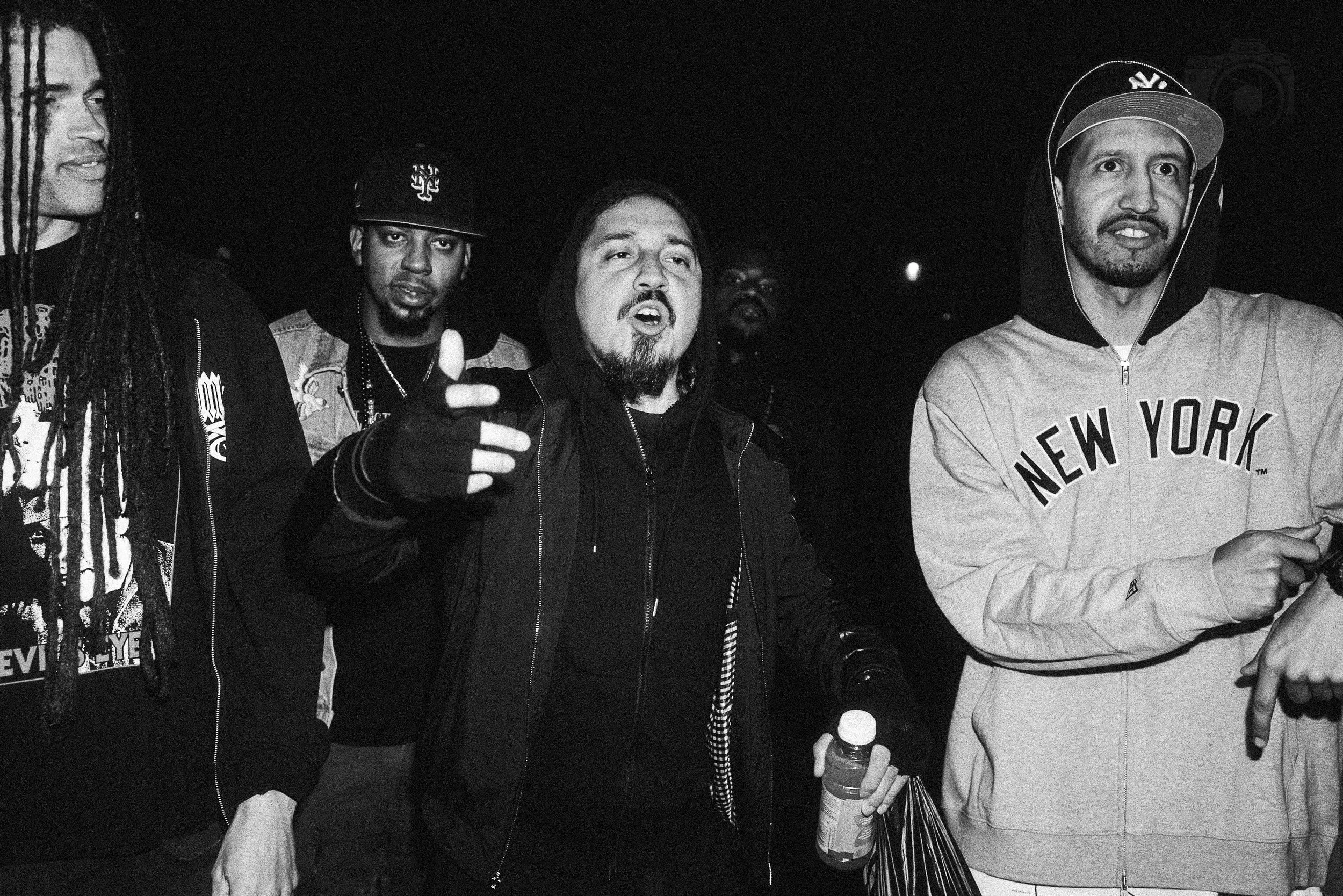 A group of five men standing outdoors at night. One man is wearing a hoodie with "New York" written on it and a cap. Another man is making a peace sign with his hand, and another is holding a water bottle and a striped bag.