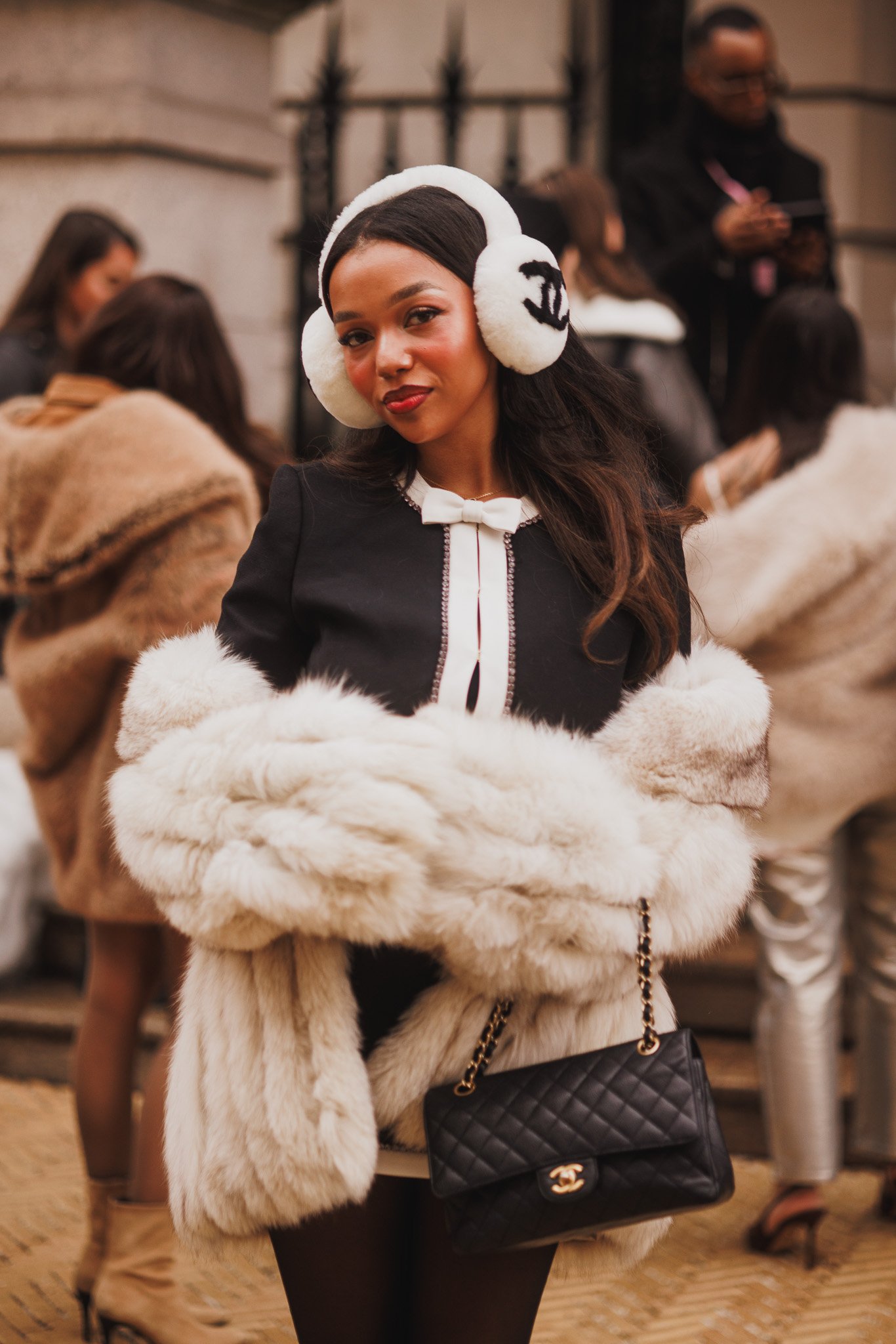 Daphne Blount wearing white earmuffs, a black dress with a white bow tie, and a white fur coat, carrying a black Chanel handbag. She is posing at an indoor social event with other people in the background.