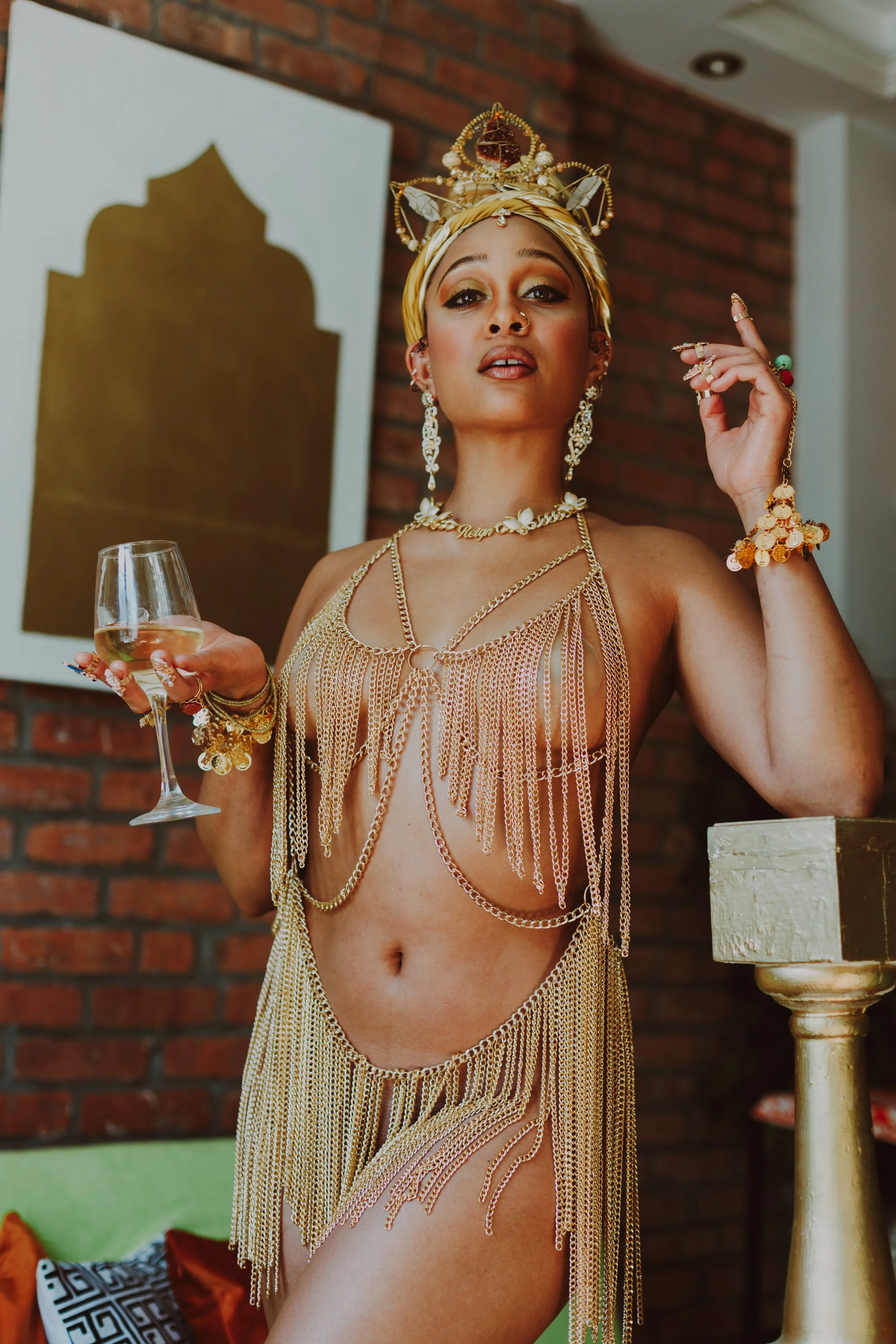 A woman wearing gold jewelry, a headpiece, and a gold chain dress, holding a glass of white wine while standing indoors with a brick wall background.