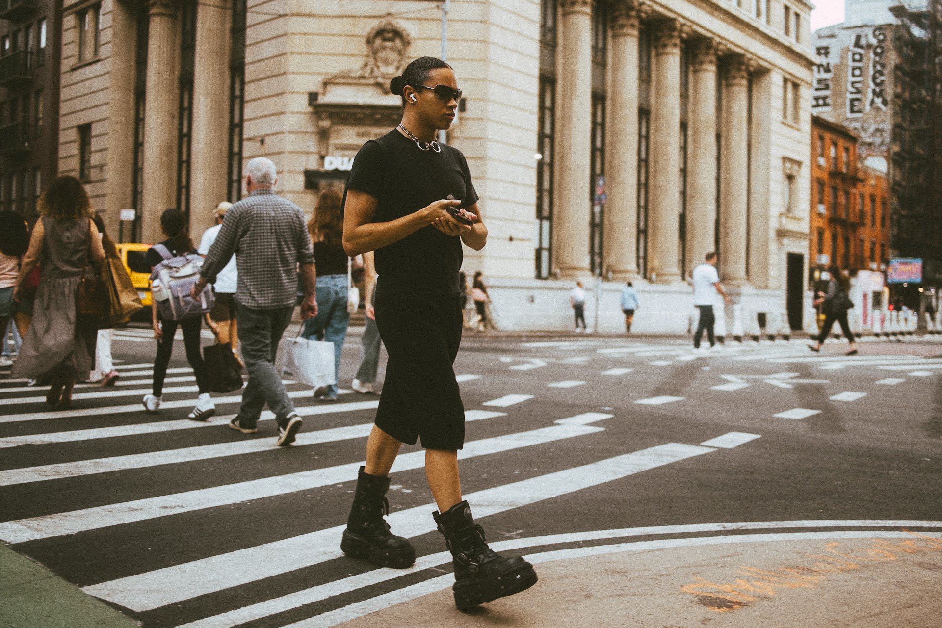 A woman with short hair, sunglasses, black t-shirt, black skirt, and punk-style chunky boots is crossing a city street on a crosswalk. She is holding a phone and wearing wireless earbuds. Several other pedestrians are crossing the street, and there i