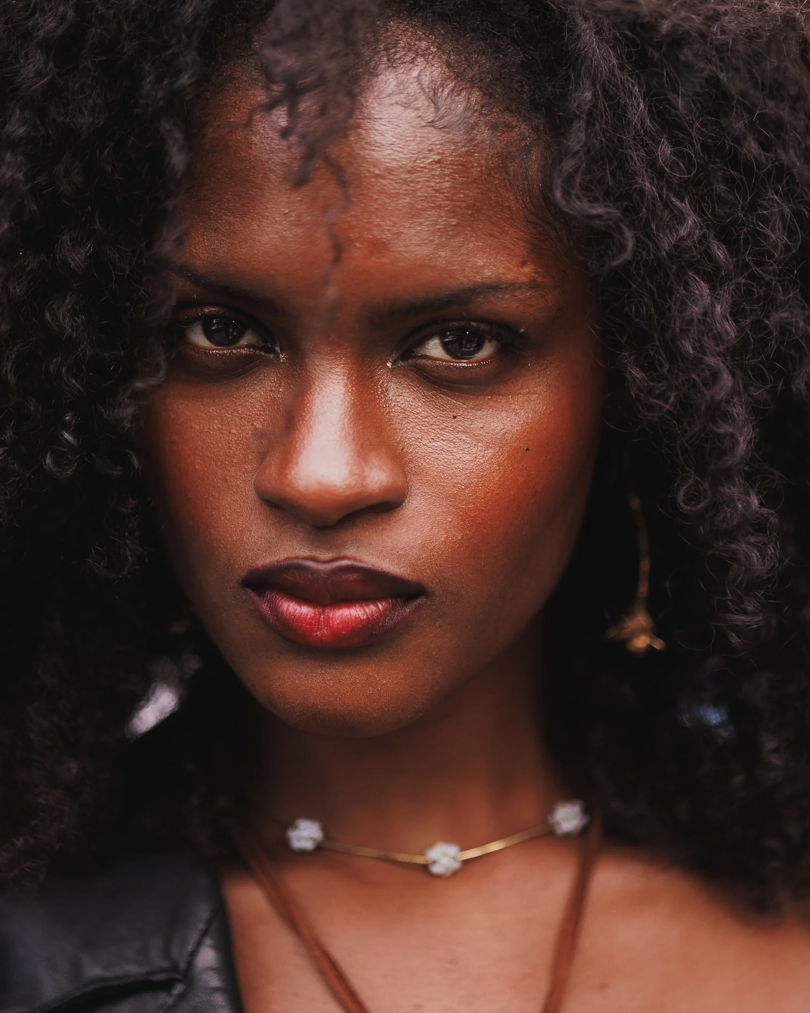 Close-up of a woman with dark curly hair, wearing a floral necklace, with part of her face and shoulders visible.