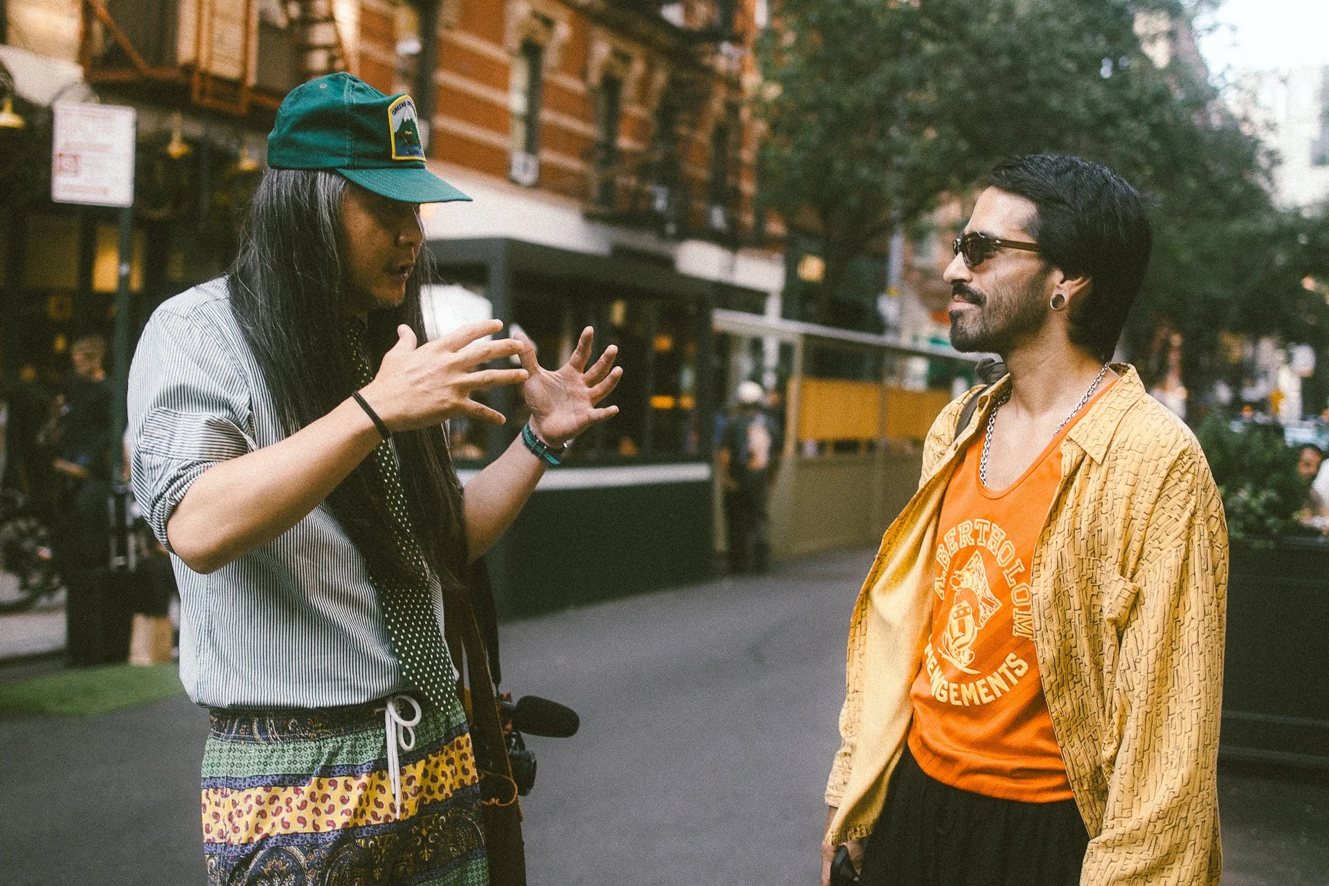 Two men having a conversation on a city street. The man on the left with long hair and a green cap gestures with his hands, the man on the right with glasses and earrings listens. Both are dressed casually, and there are buildings and trees in the ba