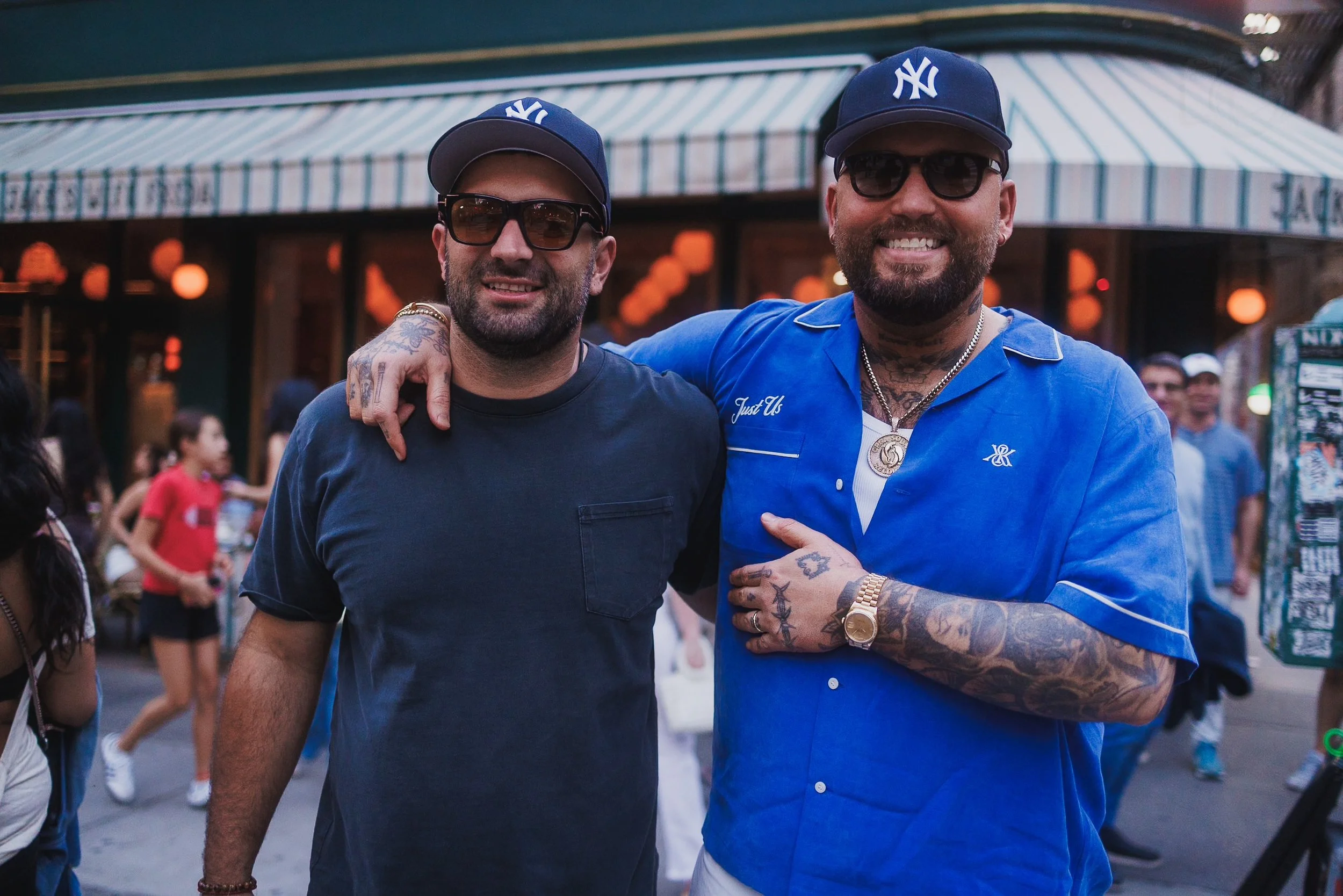 Two men wearing New York Yankees caps and sunglasses smiling, standing close together with arms around each other, in an outdoor urban setting with blurred people and a storefront in the background.