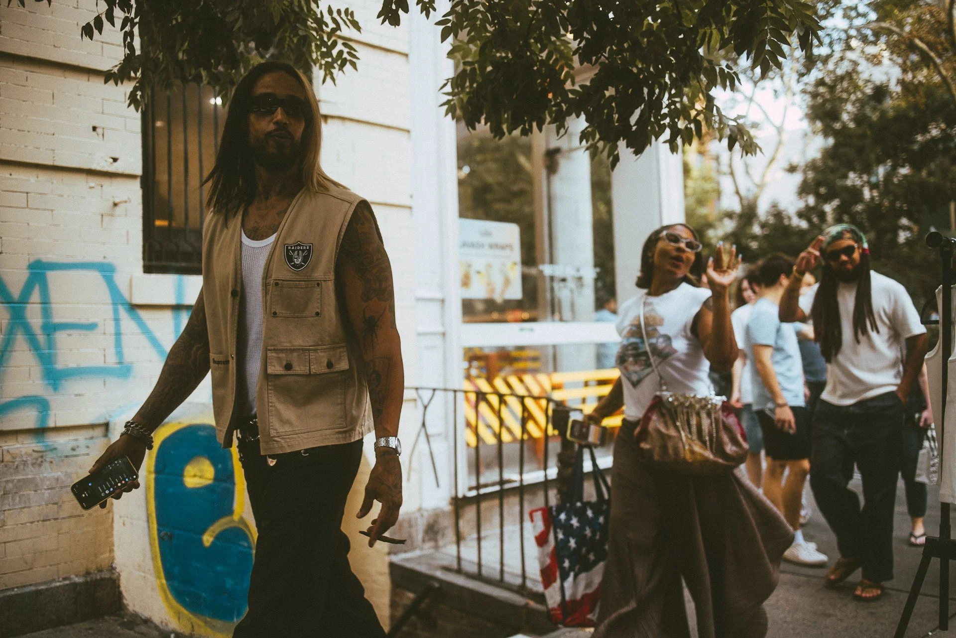 Group of people walking outside, including a man with long hair, sunglasses, tattoos, wearing a beige vest, holding a remote or electronic device, and others behind him walking with casual clothing.