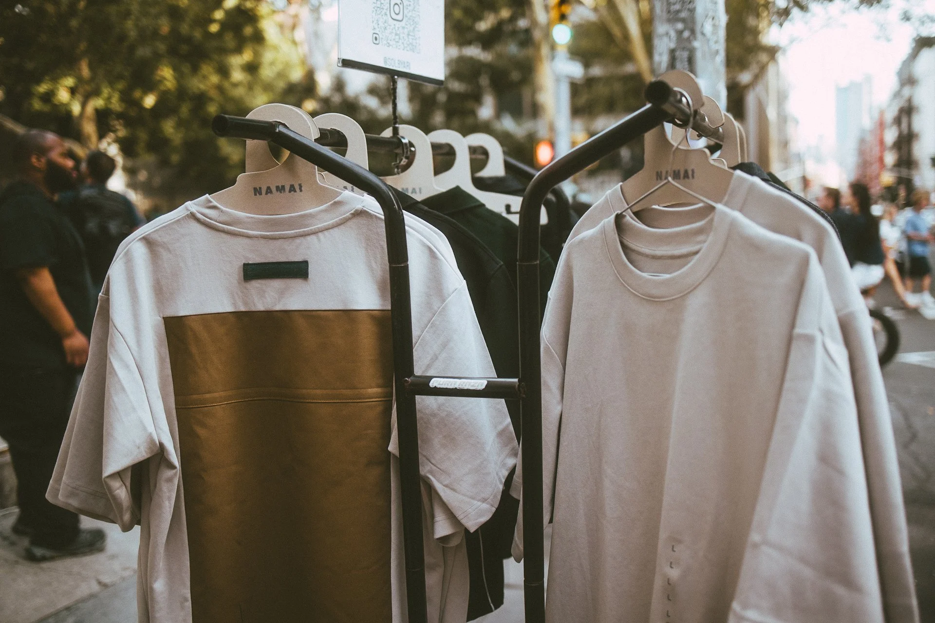 White T-shirts hanging on a clothing rack on a city street with blurred pedestrians in the background.