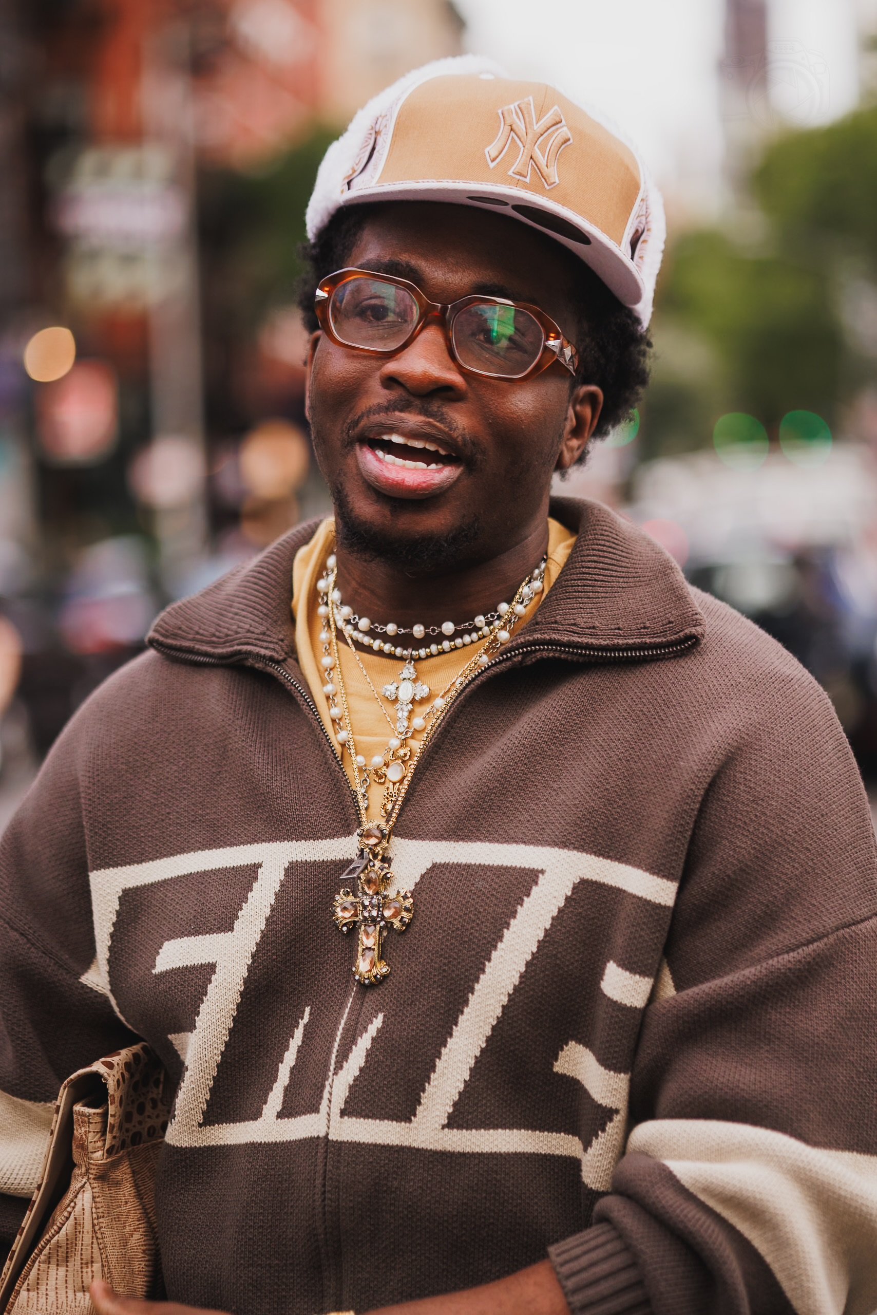 A man wearing a tan cap with a New York Yankees logo, orange-tinted glasses, a brown zip-up hoodie with large white letters, layered gold jewelry including multiple necklaces and a large cross pendant, and a beige bag, standing outdoors in an urban s