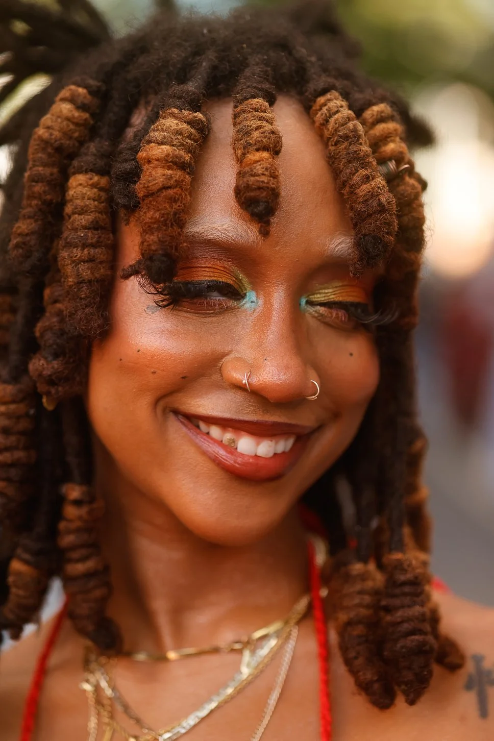Close-up of a smiling woman with colorful eye makeup, dreadlocks, nose ring, layered necklaces, and a tattoo on her shoulder.