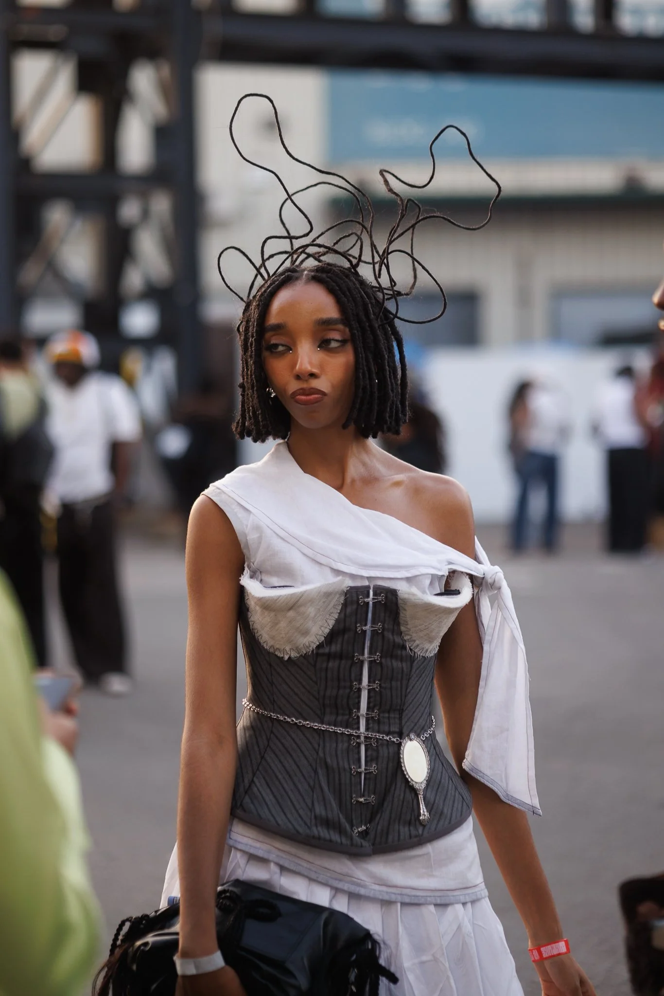 A woman with short dreadlocks and dark lipstick at an outdoor event dressed in a black and white corset with a white shirt draped over her shoulders and a white skirt, with an audience and city buildings in the background.