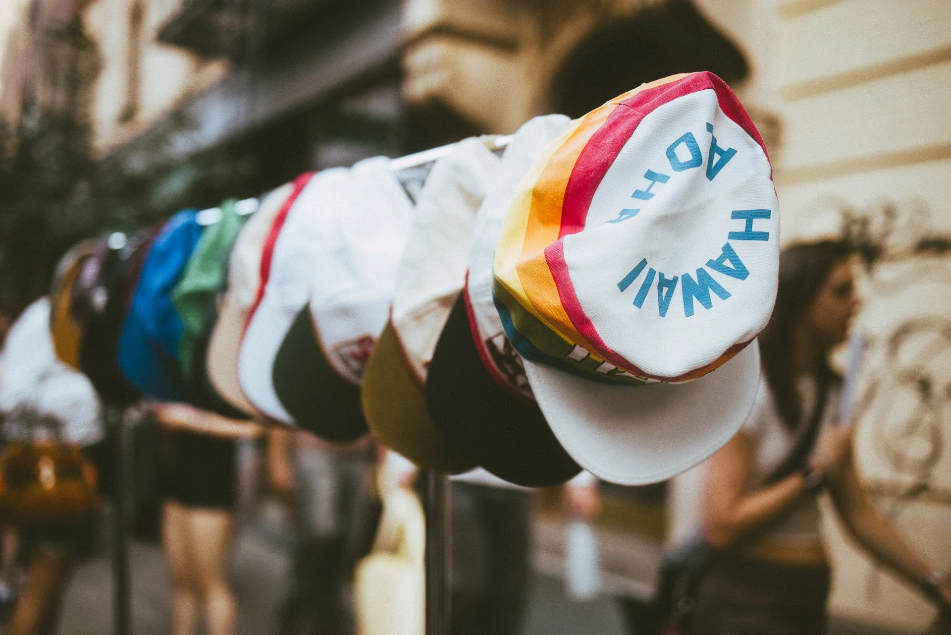 A row of colorful hats hanging on a rack in a shop, with a person blurred in the background.
