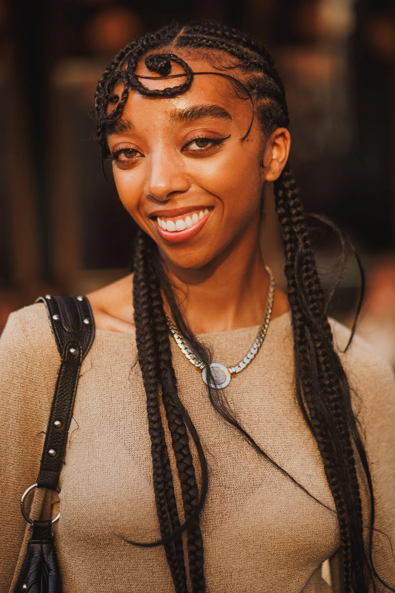 Close-up of a woman with braided hair, smiling and winking, wearing a beige top with black accents and a necklace.