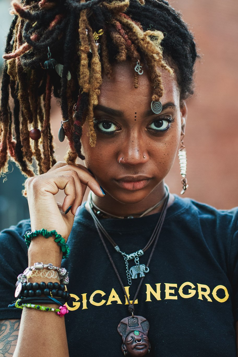 A woman with dreadlocks and intense gaze, wearing multiple necklaces with cultural pendant charms, colorful bracelets, and a black T-shirt with the words 'Magica Negra.'