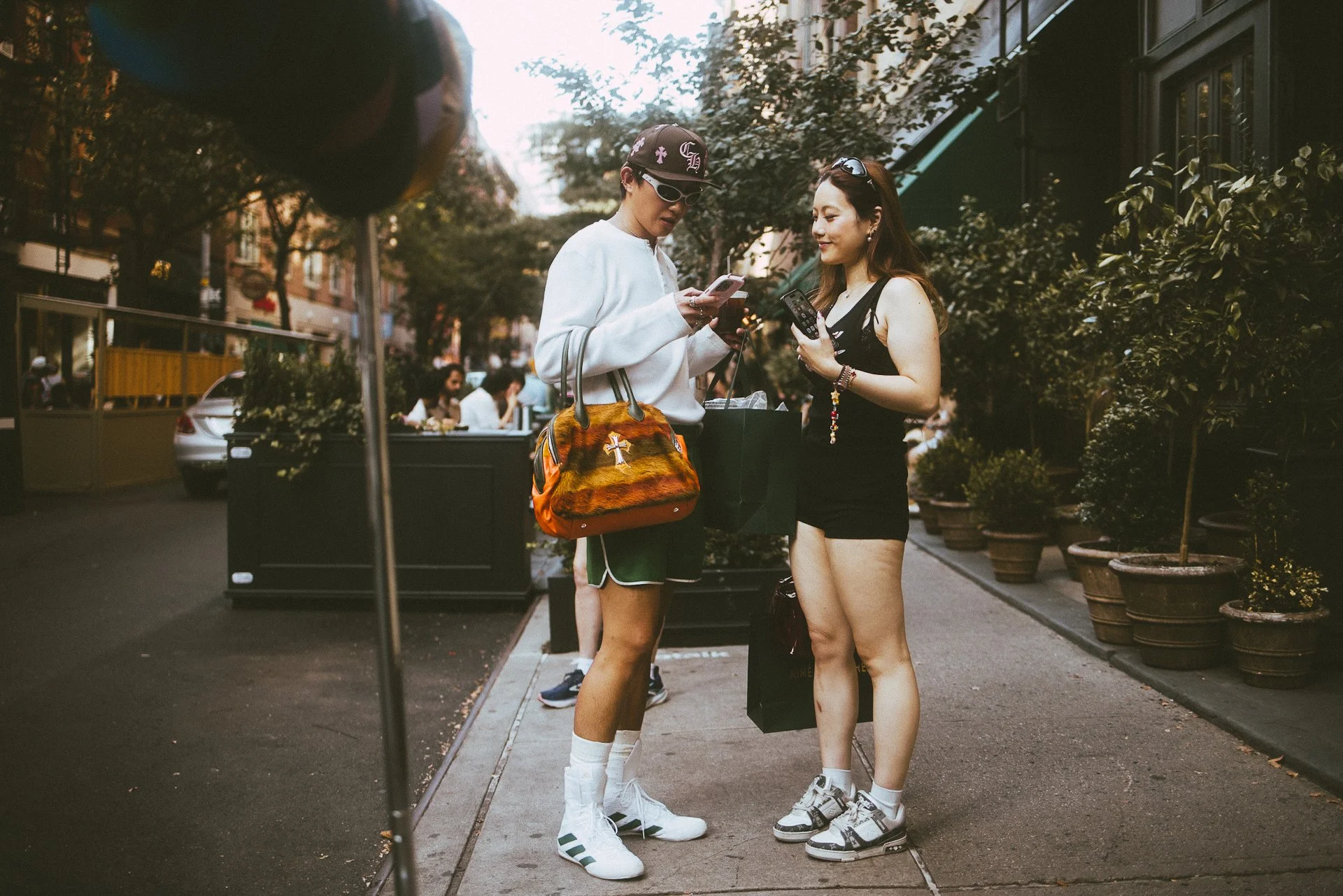 Two women standing on a city sidewalk, looking at their phones, with potted plants and buildings in the background.