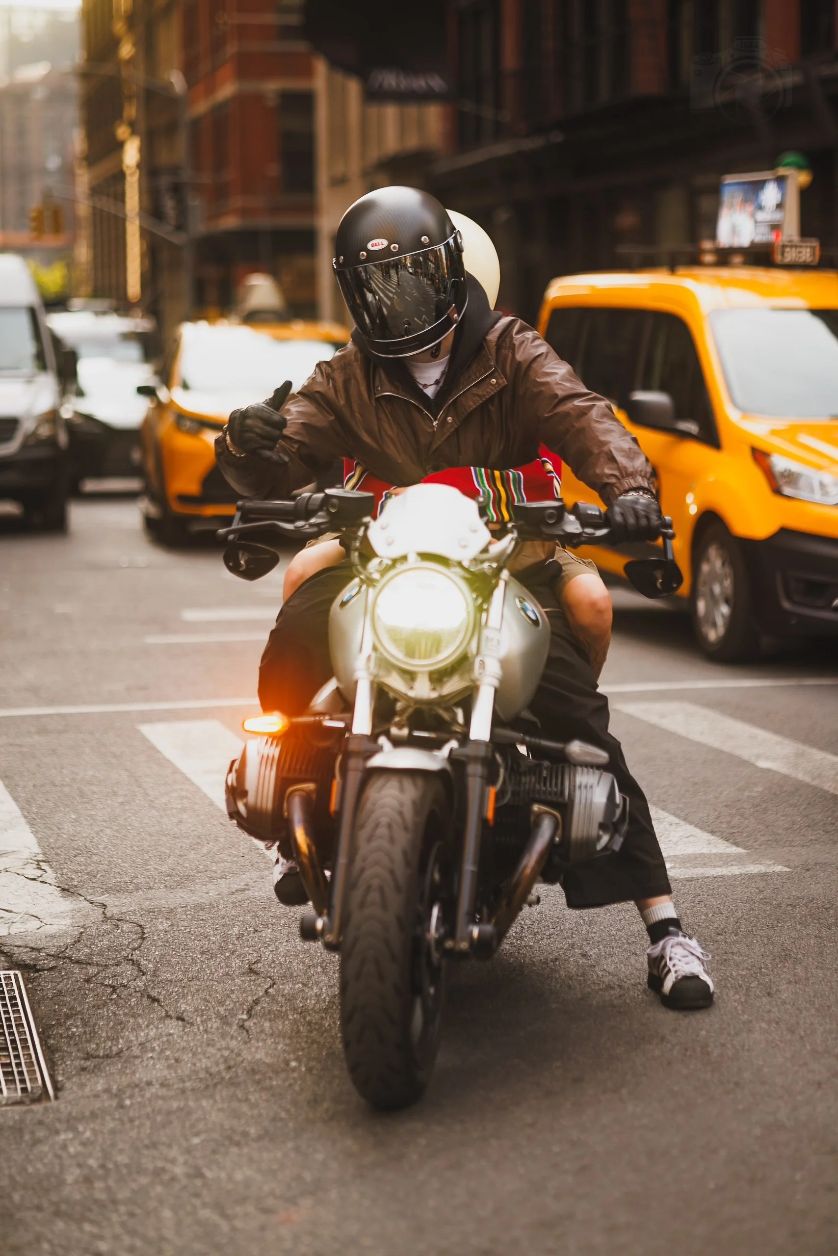 A person riding a motorcycle in an urban street, wearing a black helmet with a tinted visor, a brown jacket, and black shorts. The person is giving a thumbs-up, and yellow taxis and cars are visible in the background.