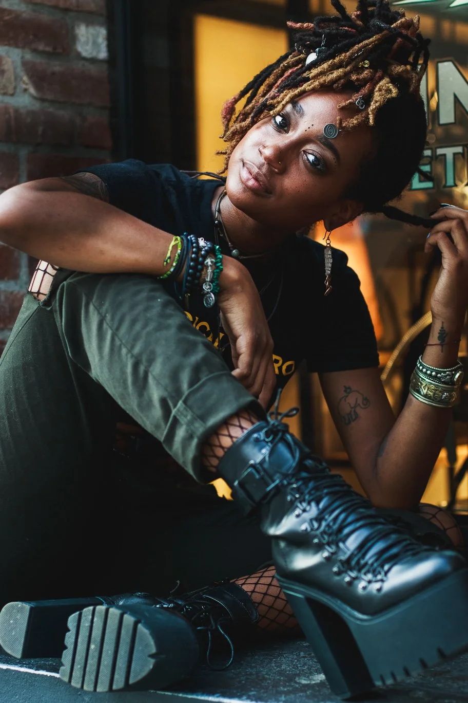 A young woman with dreadlocks and tattoos sitting against a brick wall, wearing black combat boots, cargo pants, and a black T-shirt, accessorized with bracelets, necklaces, and earrings.