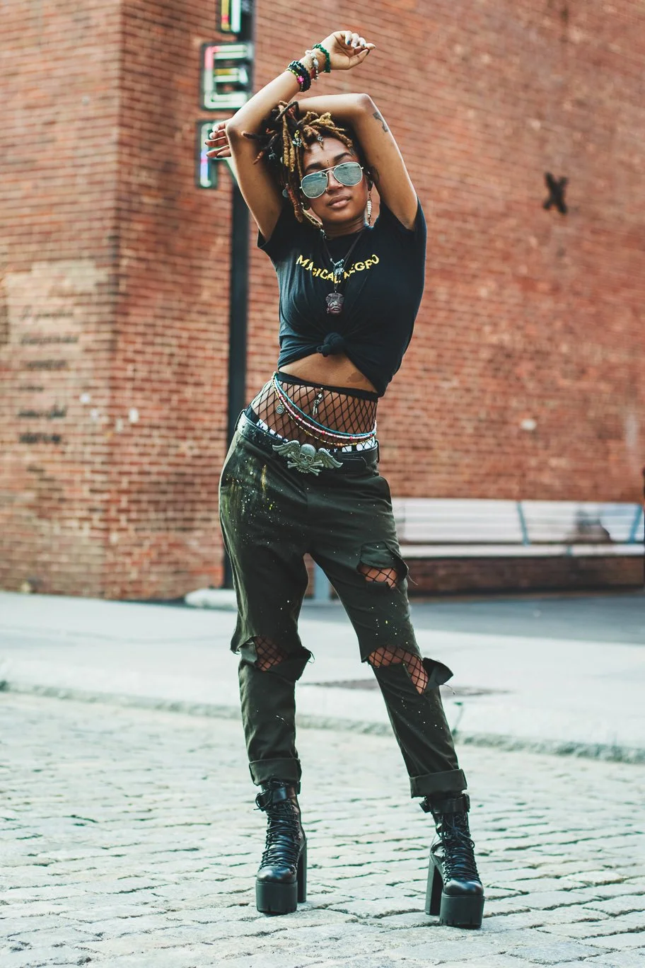A woman with dreadlocks and sunglasses striking a pose outdoors in front of a brick building, wearing a black knotted t-shirt, ripped black pants, and platform boots.