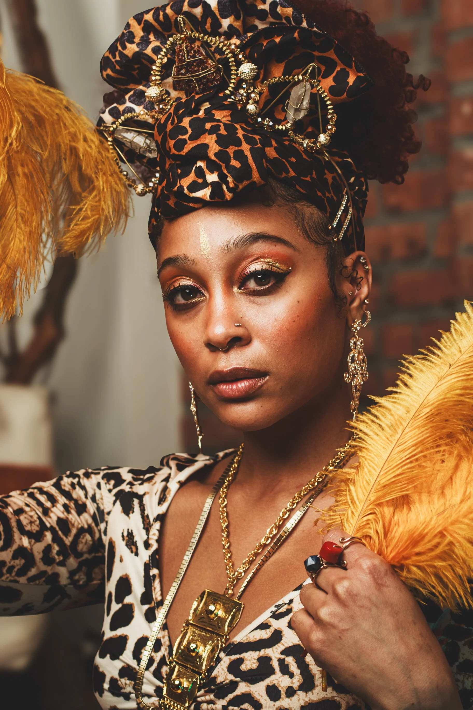 Woman with leopard print headscarf, gold jewelry, and makeup, holding a yellow feather, with a brick wall in background.