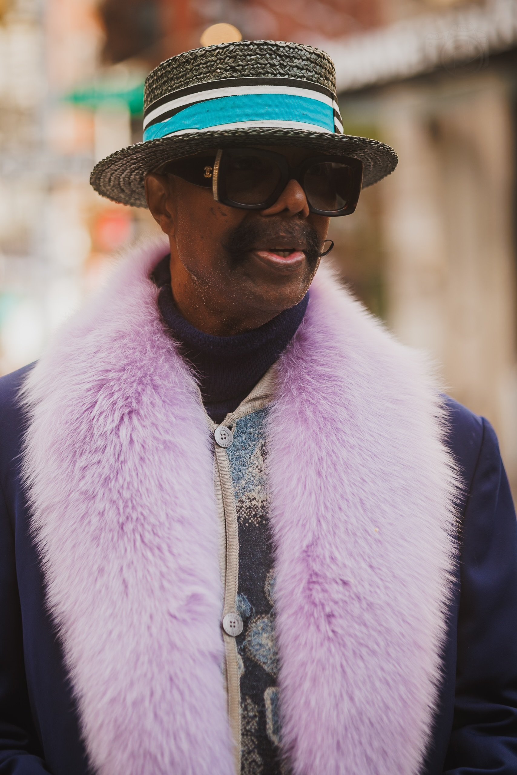 A man wearing a black woven hat with a striped blue, white, and black band, dark sunglasses, a dark blue coat with a pink fur collar, and a patterned shirt underneath.