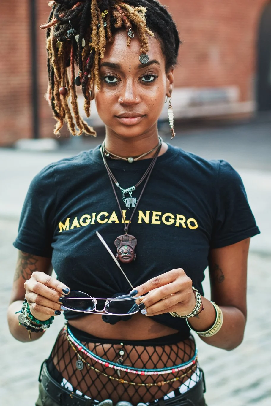 A young Black woman with dreadlocks and facial piercings, wearing a black T-shirt with the words 'Magical Negro,' holding sunglasses, standing outdoors with a blurred brick building background.