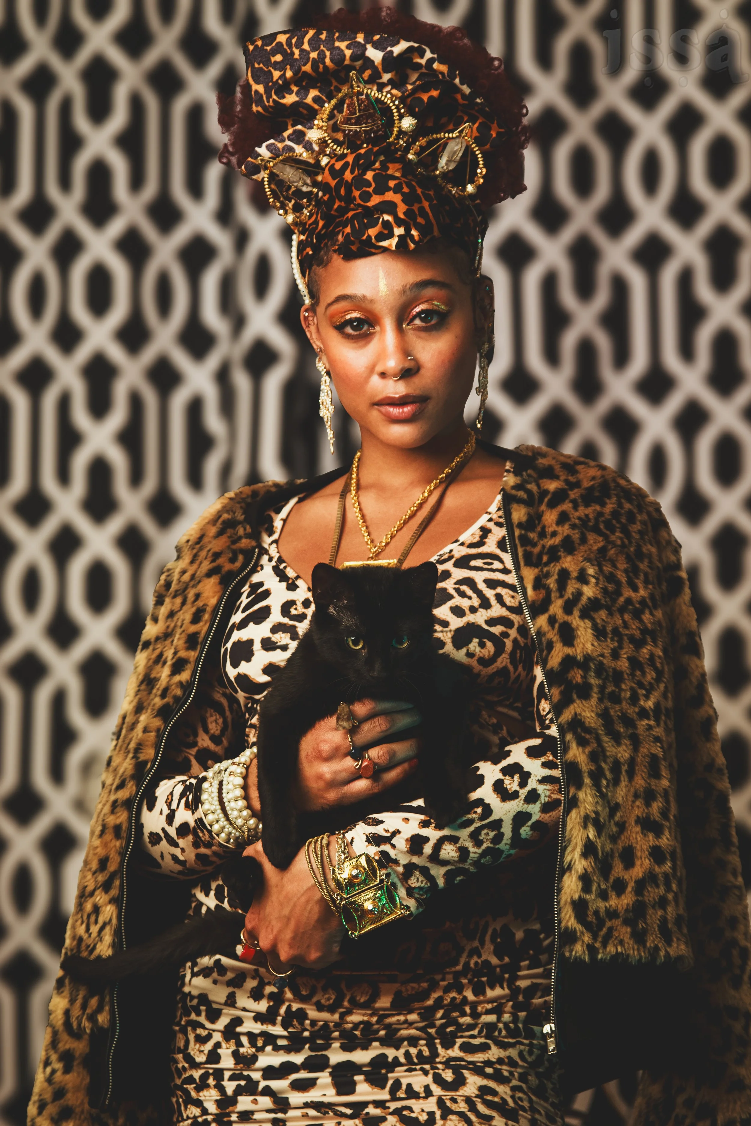 A woman with dark skin and elaborate jewelry, wearing a leopard print headscarf, leopard print dress, and a leopard print jacket. She holds a black cat and stands against a decorative black-and-white patterned background.