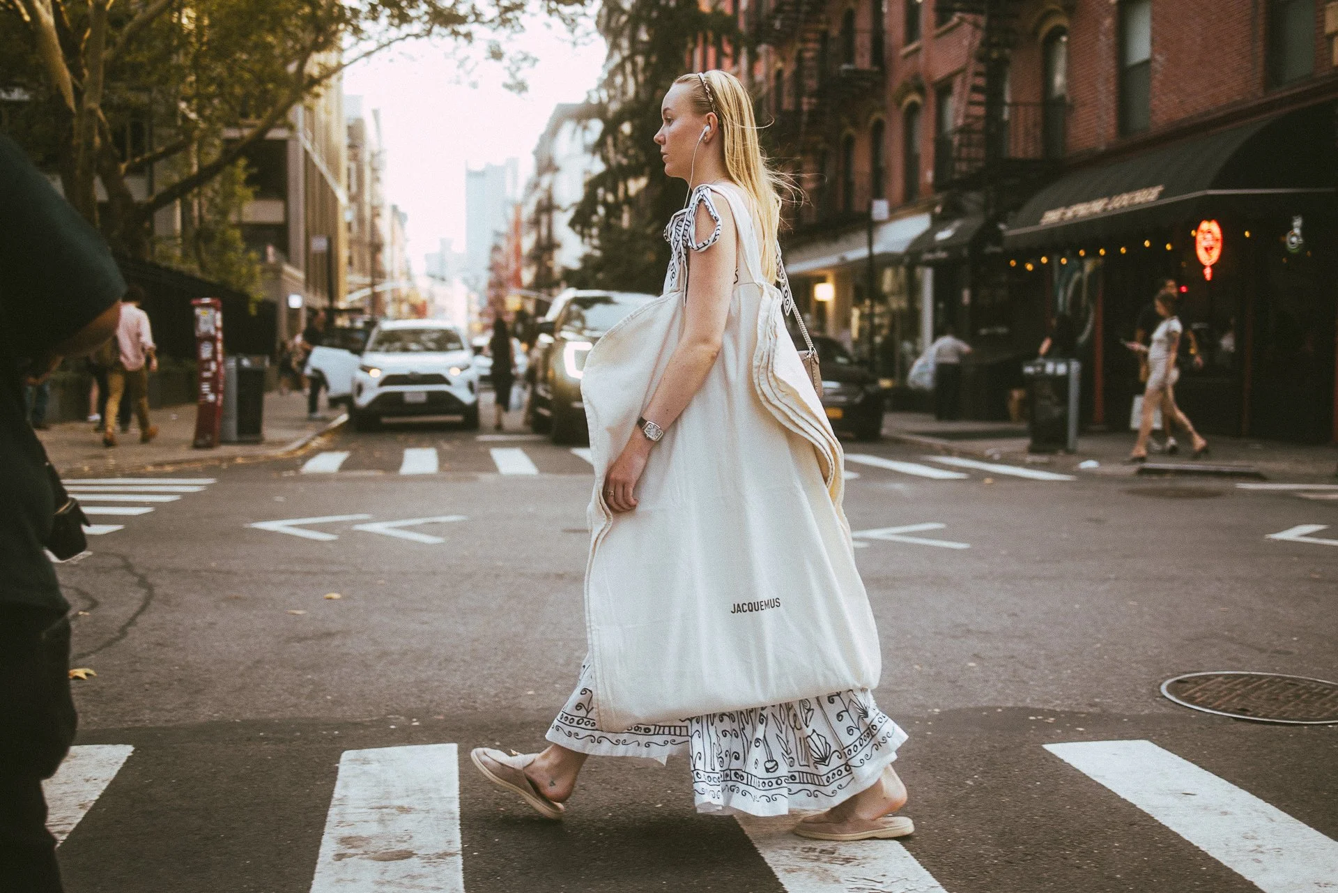 A woman crossing the street in an urban area, holding a large white shopping bag with 'JACQUEMUS' printed on it, wearing a light-colored dress with a patterned skirt, sneakers, and earphones.