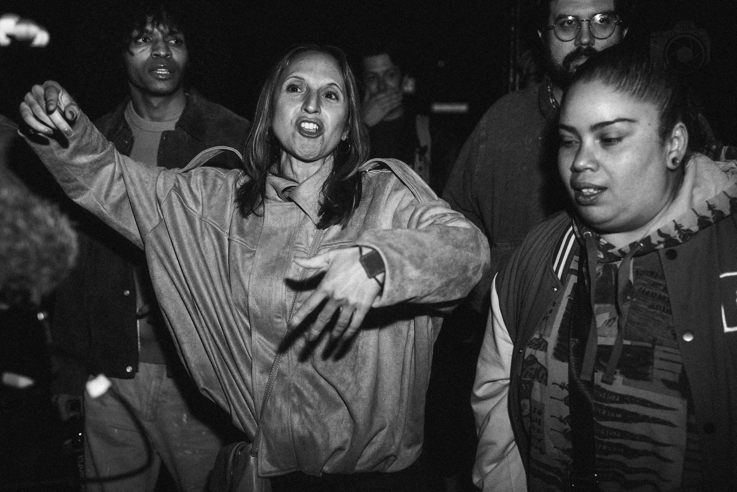 A group of four people gathered together in a dark setting. The woman in the center appears to be speaking or singing passionately, with an expressive face and arm raised. The others around her seem attentive or engaged, with one person in the backgr