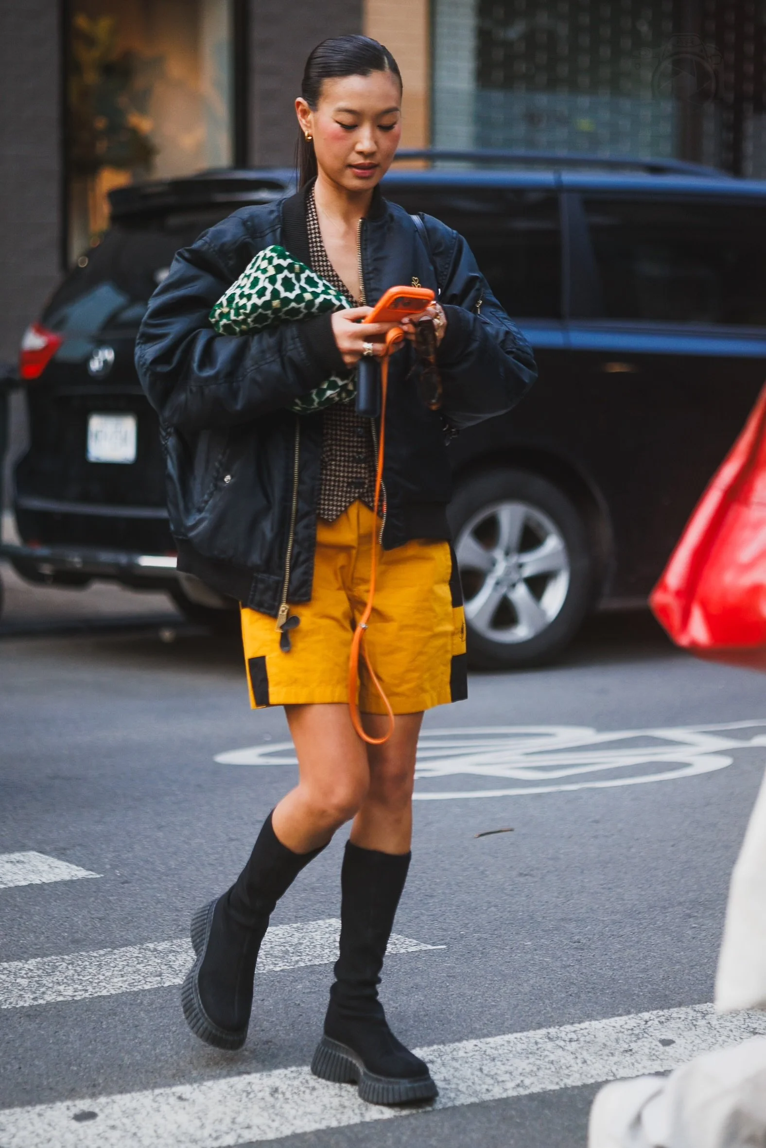 A young woman wearing a black jacket, yellow shorts, and black knee-high boots walking on a crosswalk while looking at her phone, carrying a green patterned bag.