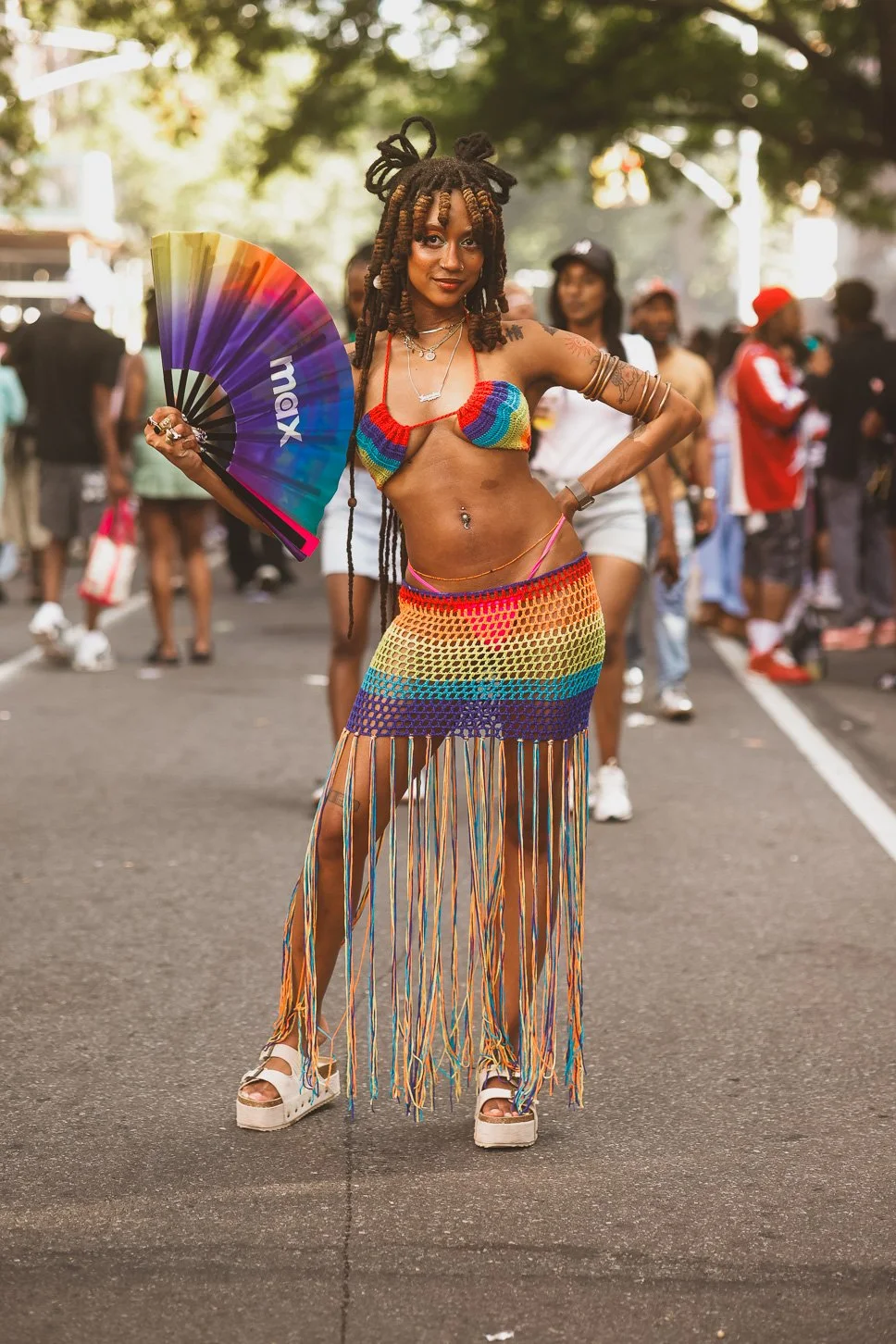 A woman in colorful crochet festival outfit holding a rainbow fan at an outdoor event.