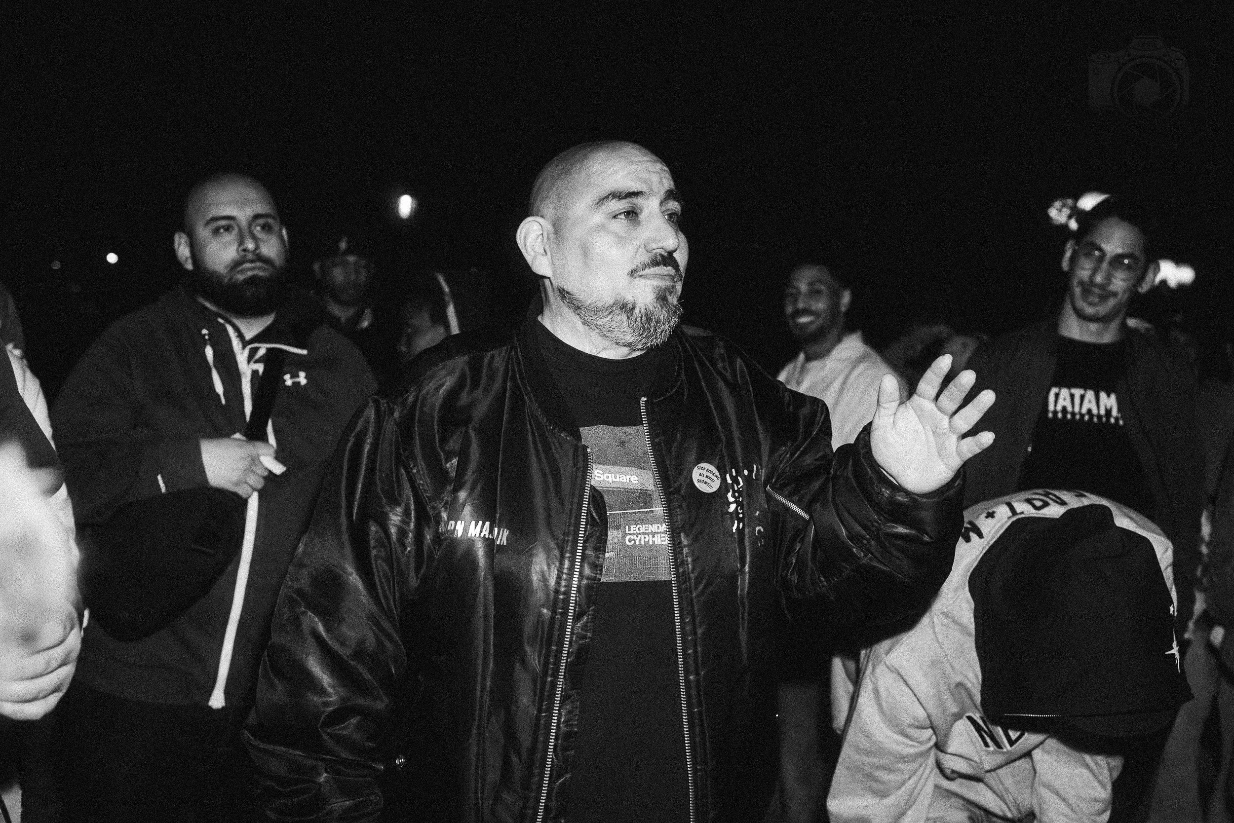 A group of people gathered outdoors at night, with one man in the center wearing a leather jacket and gesturing with his hand, while others around him look on.