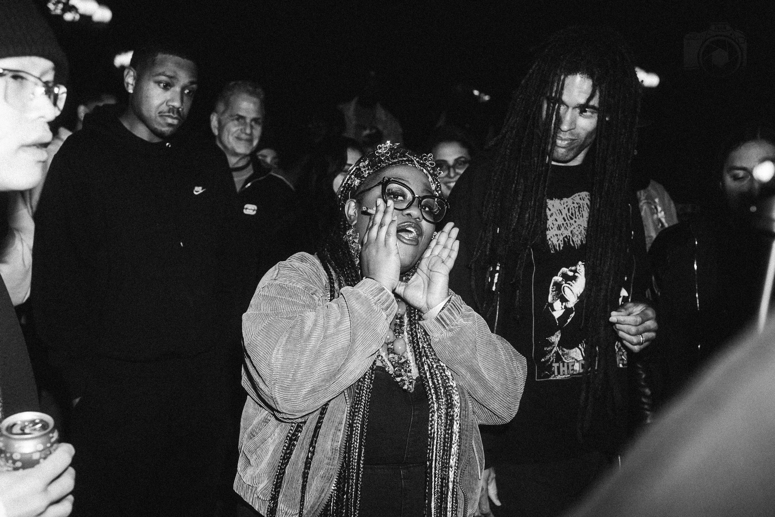 A black and white photo of a group of people at a social gathering, including a woman with glasses and braids in the foreground, appearing to be surprised or excited, with others smiling and chatting around her.