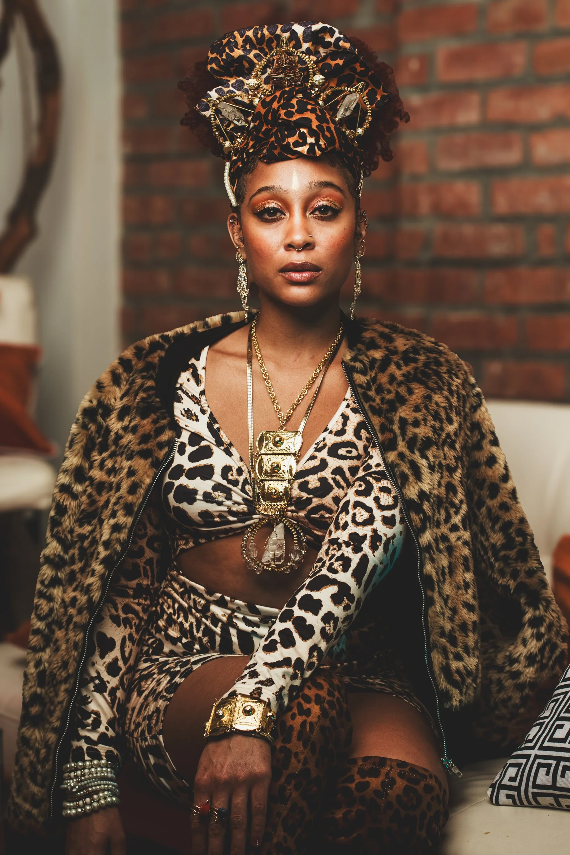A woman with dark skin, wearing leopard print clothing, big animal print headwrap adorned with jewelry, and multiple gold and pearl accessories, sitting on a couch with a brick wall background.