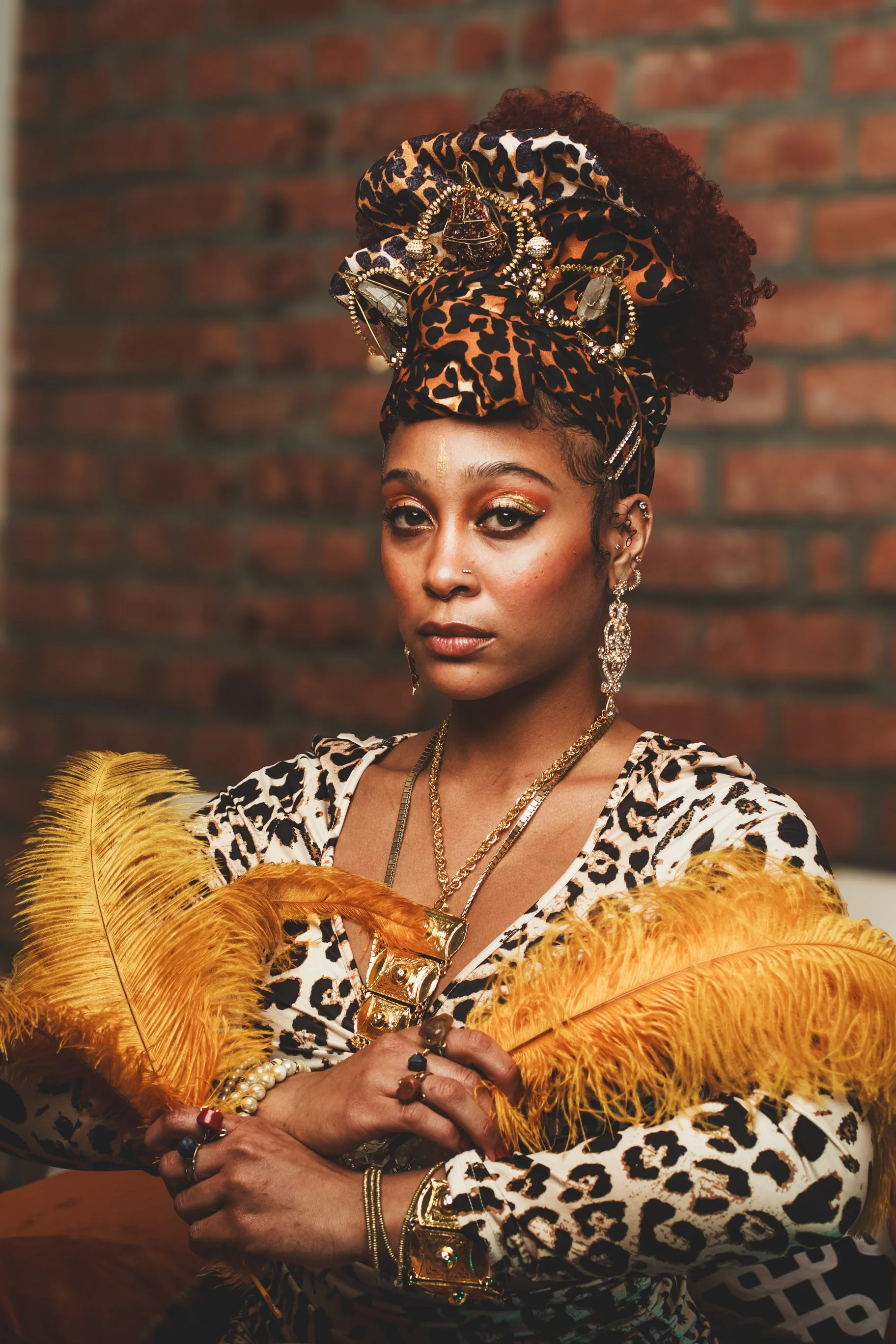 A woman with dark skin in leopard print attire, adorned with gold jewelry, and wearing a leopard print headwrap decorated with jewelry, standing against a brick wall background.
