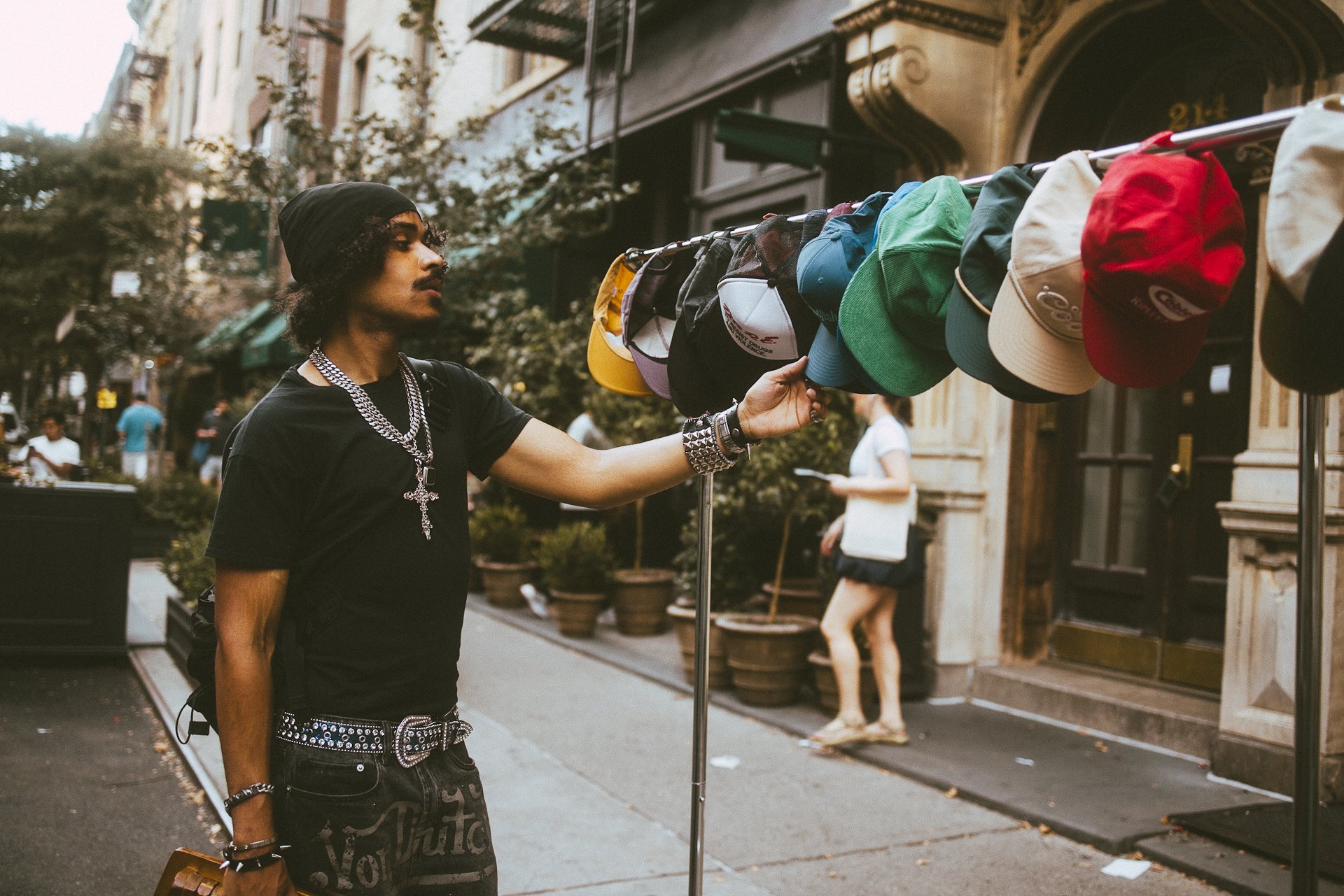 A person with dark curly hair, wearing a black head covering, black shirt, and jewelry, stands on a city sidewalk, reaching towards a display of colorful caps on a rack.