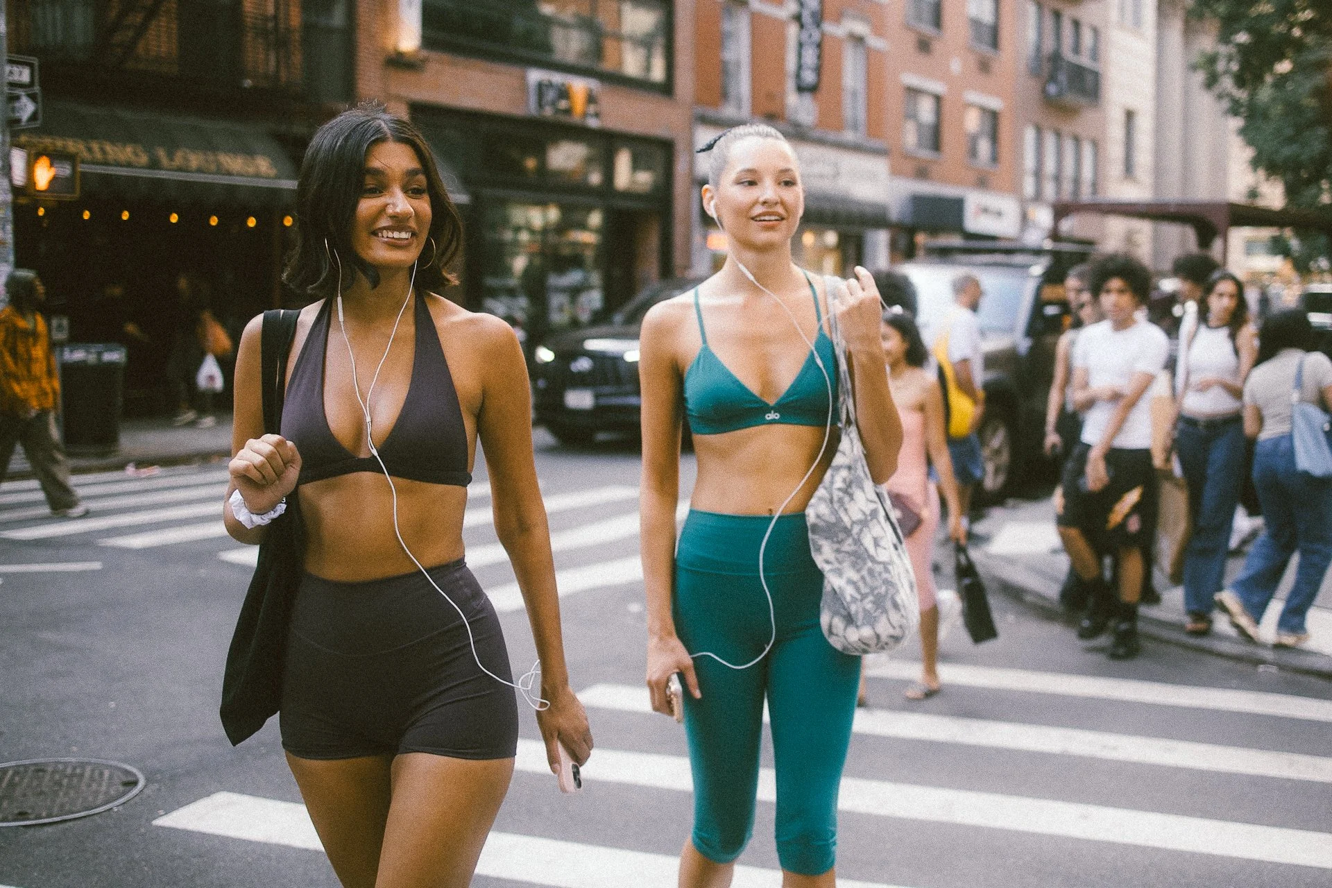 Two young women walking together across a city street while listening to music with earbuds. One is wearing a black sports bra and shorts, and the other is in a teal sports bra and leggings, carrying a patterned tote bag.