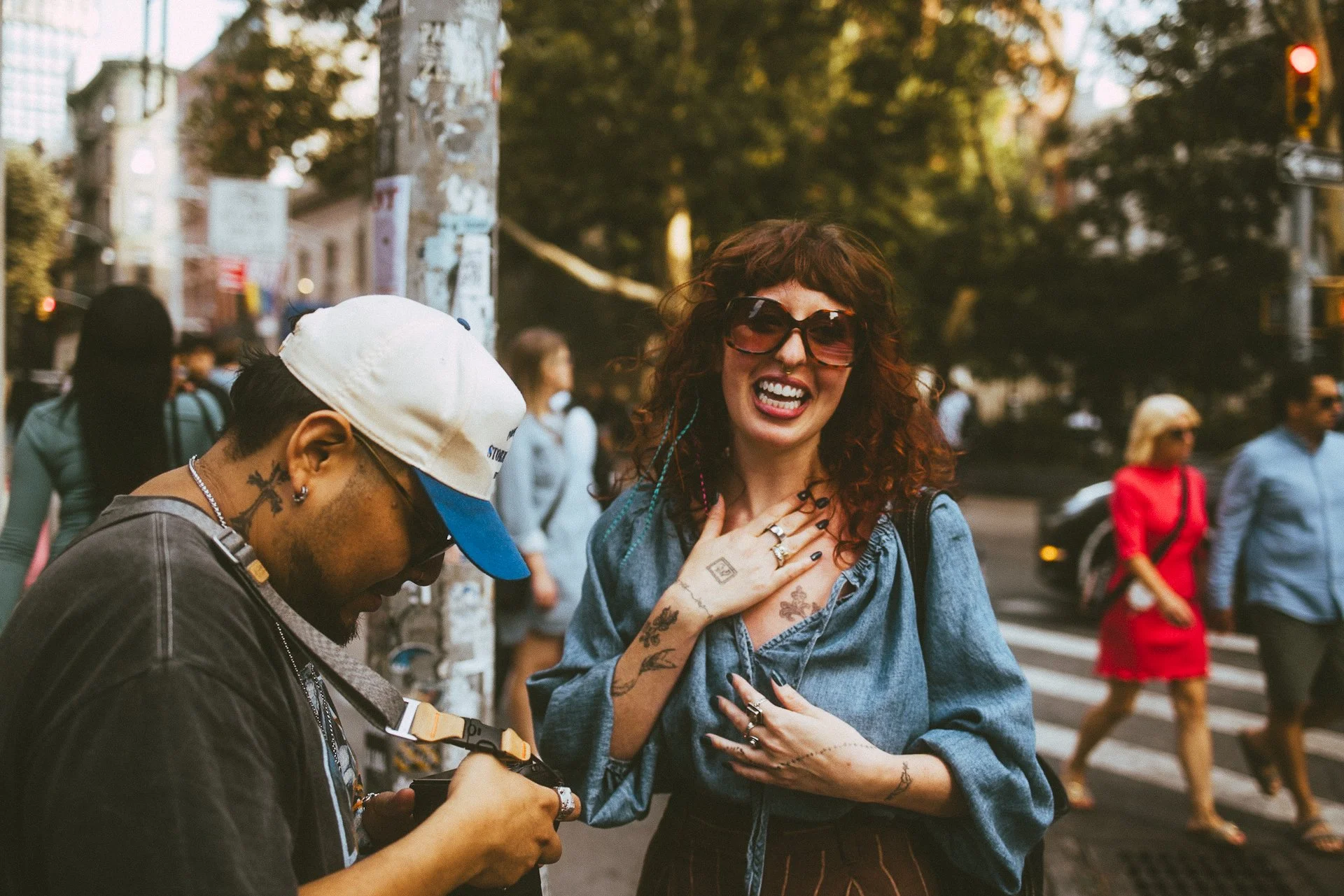 A woman with curly hair, wearing sunglasses and a denim jacket, smiling and touching her chest, interacting with a man wearing a white cap and glasses on a city street. People are walking in the background.