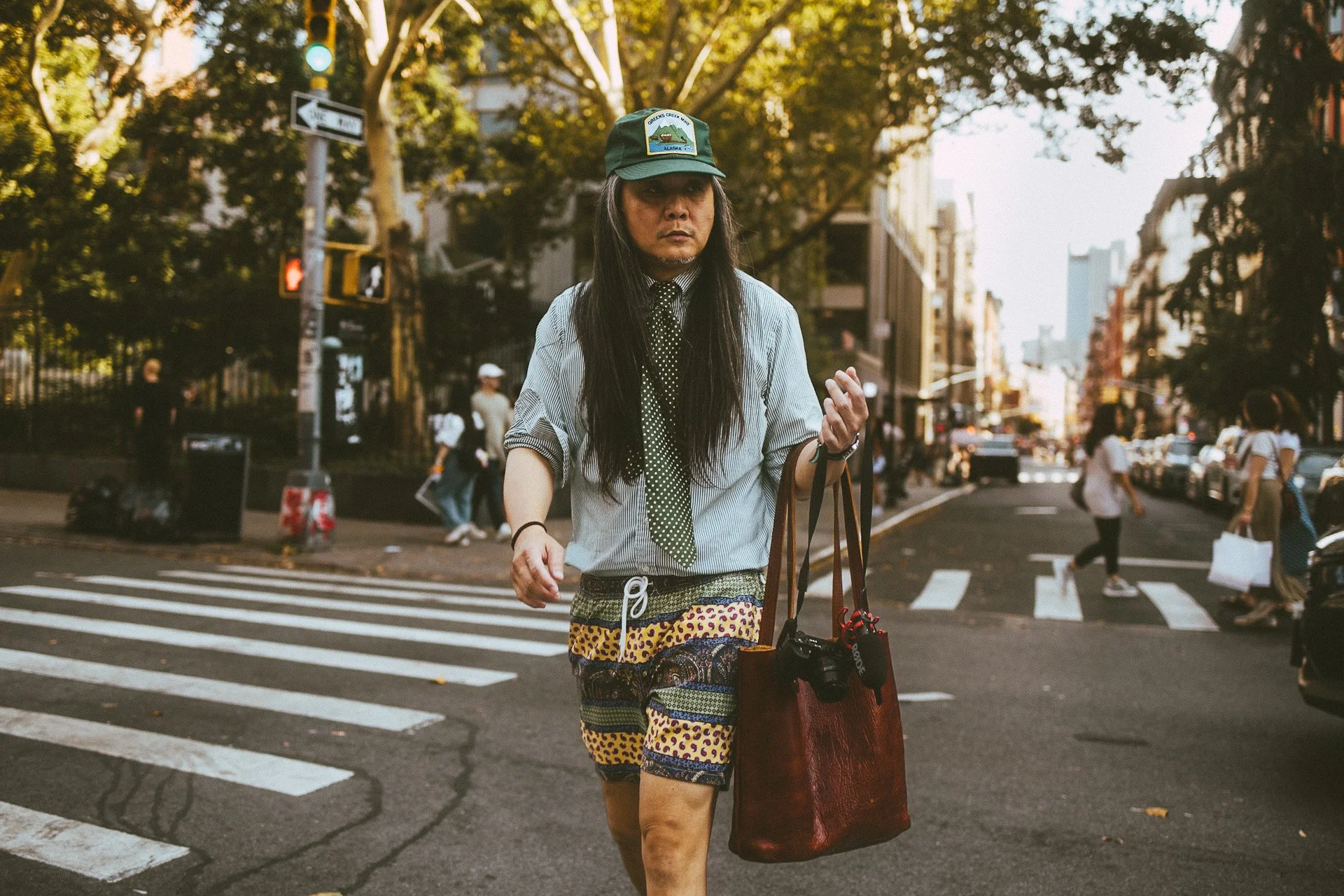 A woman wearing a green cap, a striped shirt, polka dot tie, and colorful shorts crosswalks in an urban street with other pedestrians and buildings in the background.