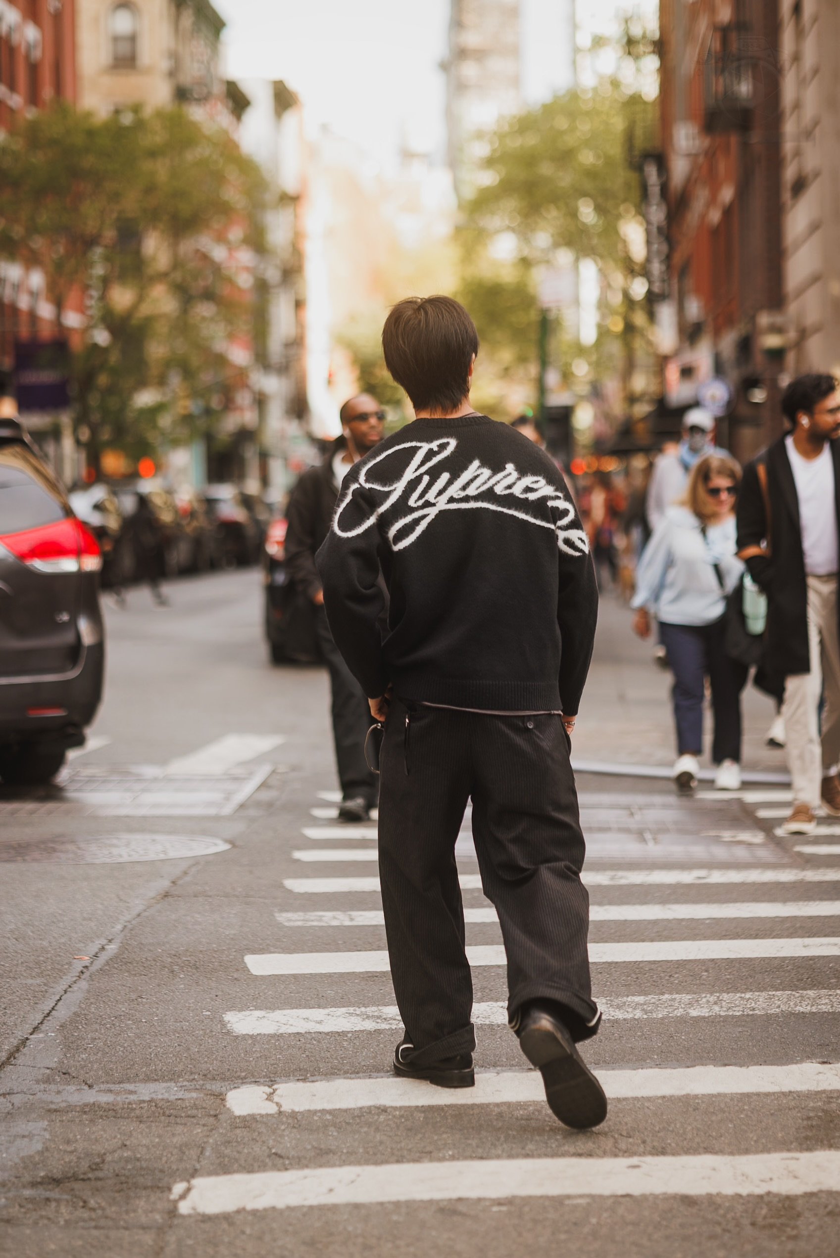 A person with short dark hair wearing a black sweater with the word 'Supreme' written on the back, walking across a crosswalk on a city street surrounded by pedestrians and parked cars.