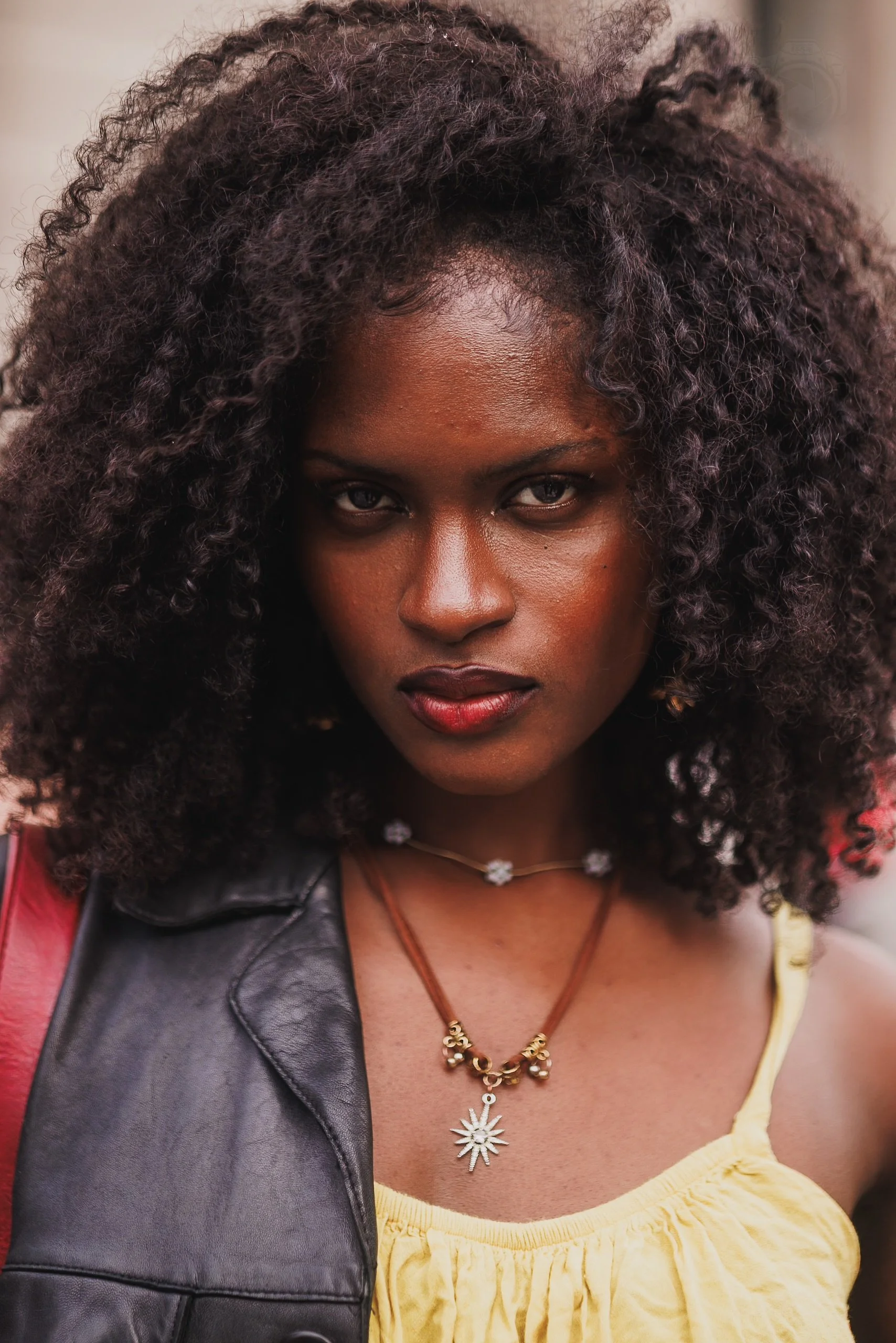Close-up of a woman with dark curly hair, wearing layered necklaces and a yellow top with a black leather vest.
