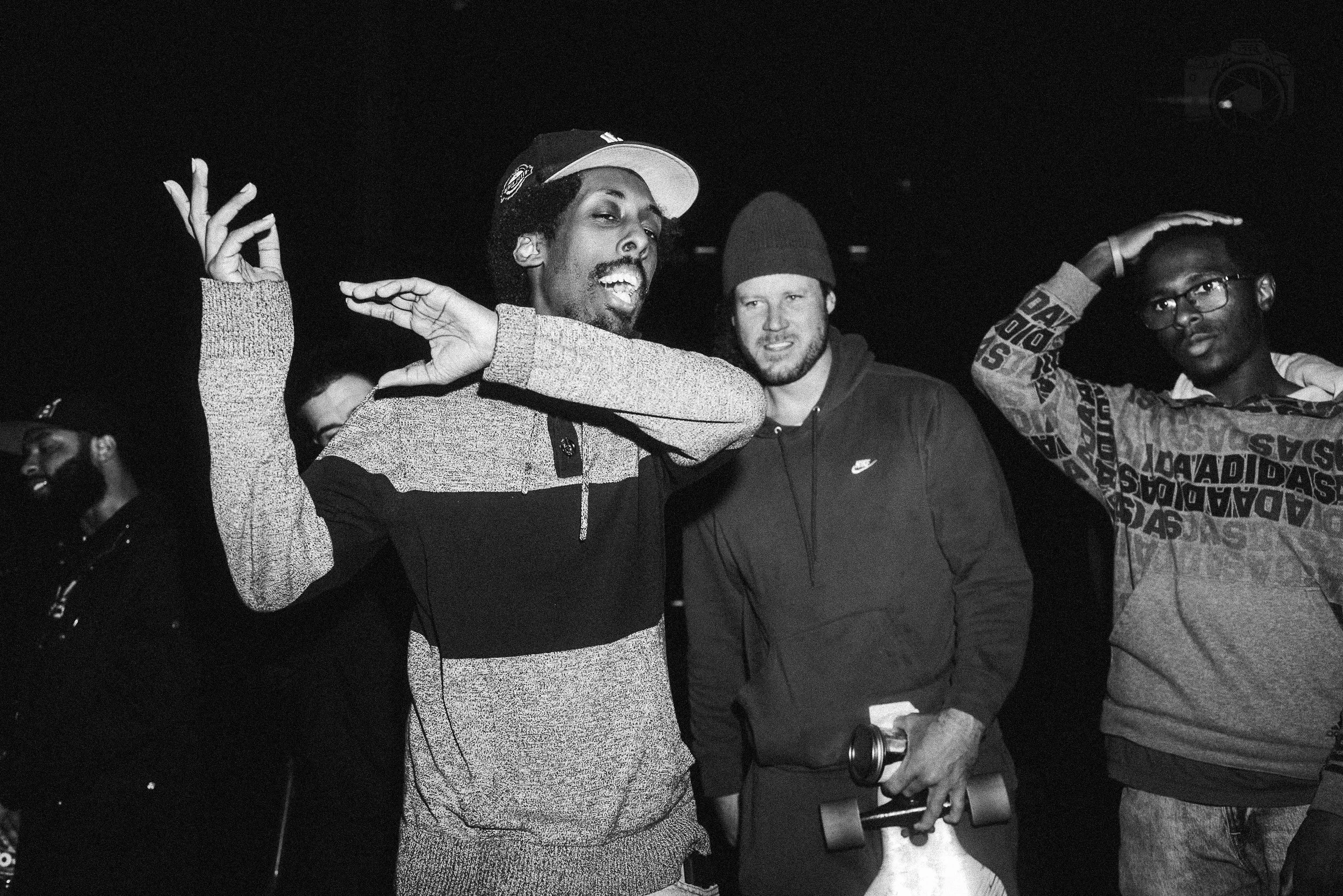 A group of young men gathered together, with two of them making hand gestures and one holding a beverage, in a dark indoor setting.