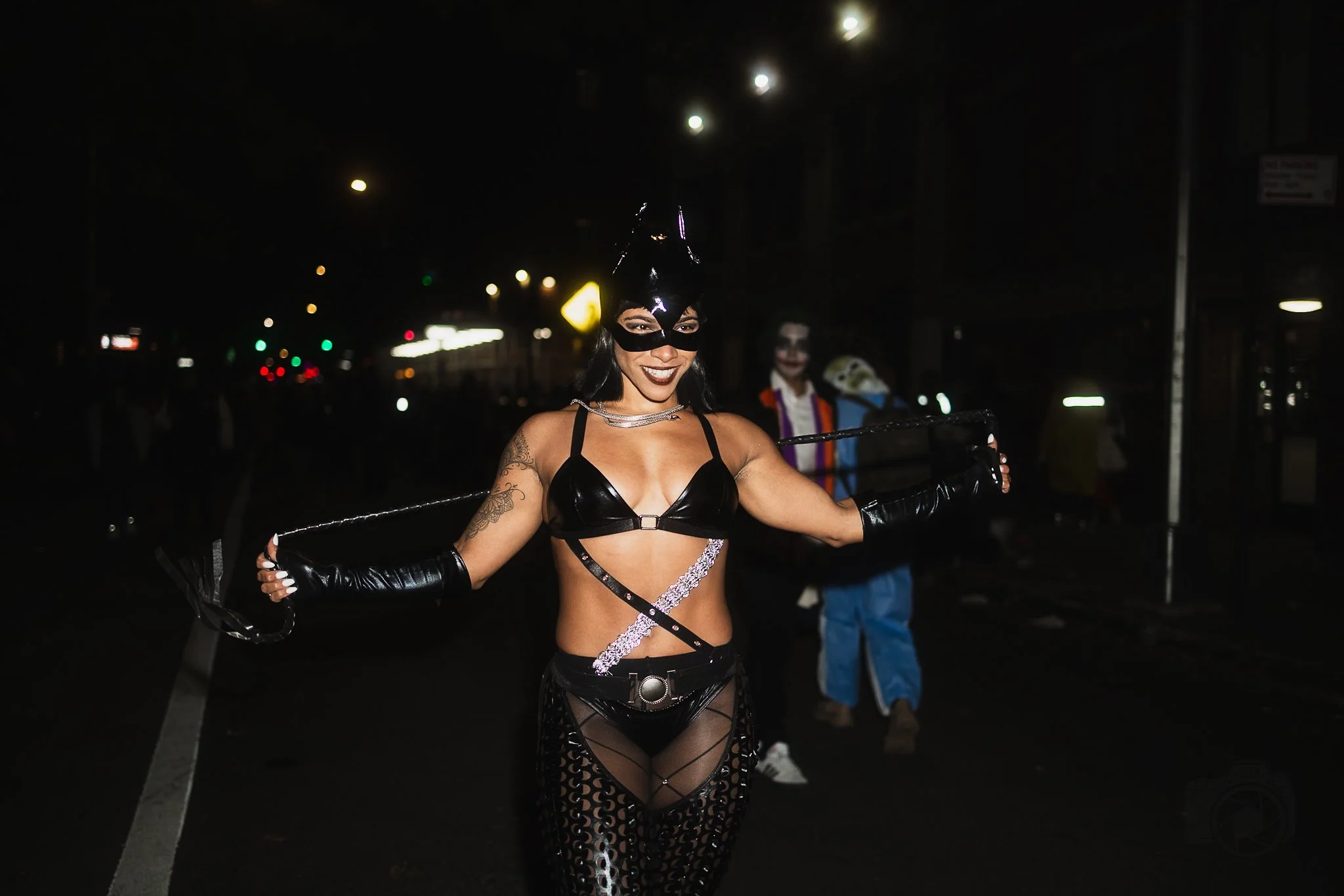 Person dressed as Catwoman in black costume with a whip, smiling at night, with a clown in costume in the background.