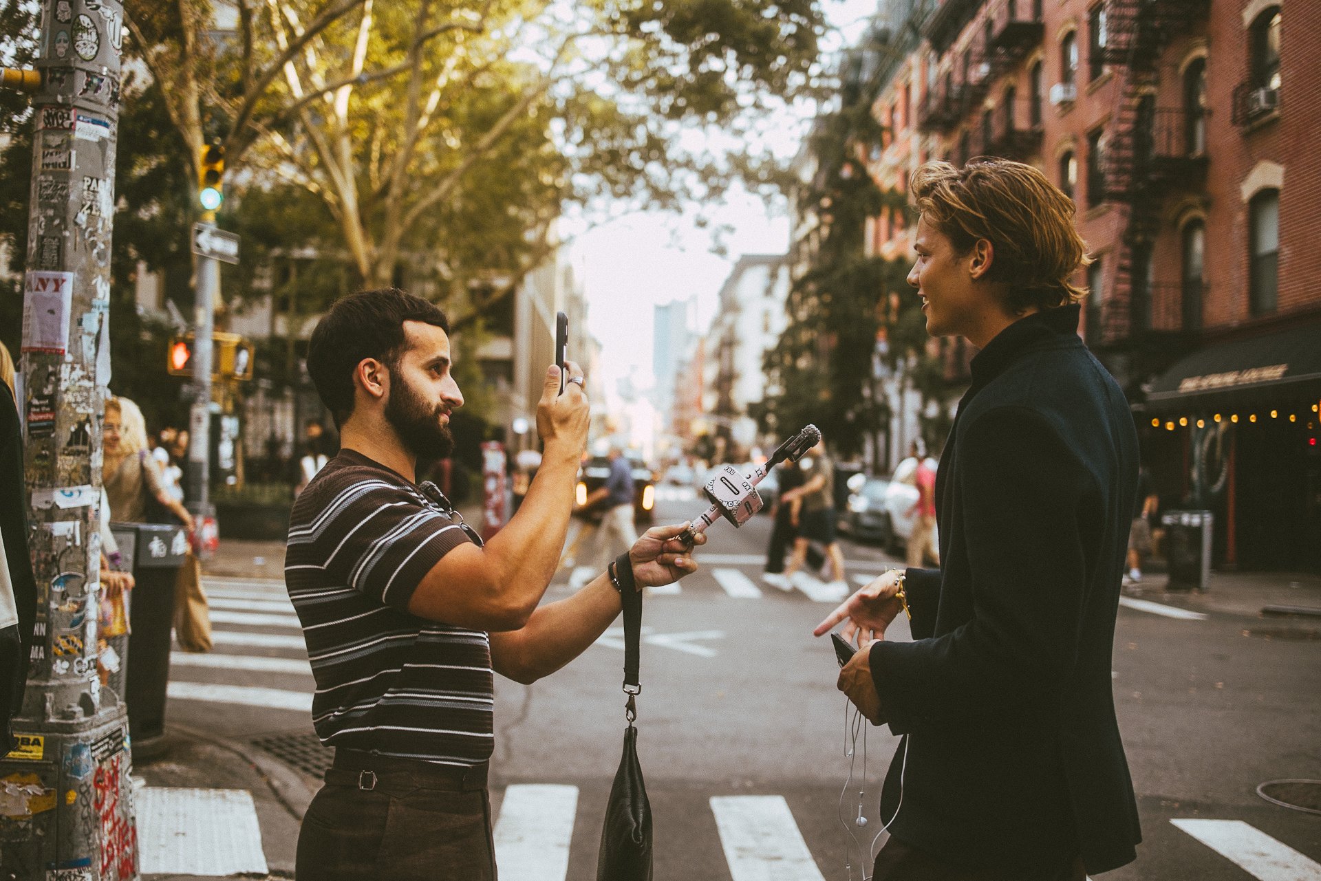 Man with beard and dark striped shirt interviewing a woman with short hair and black blazer on a city sidewalk.