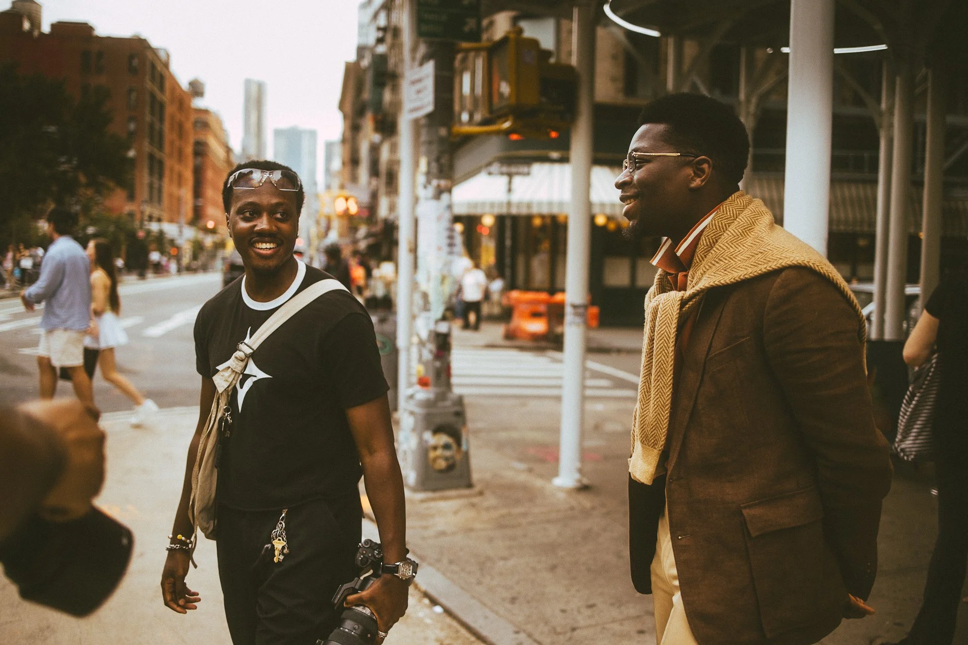 Two young men are smiling and talking on a city street, with pedestrians and city buildings in the background.