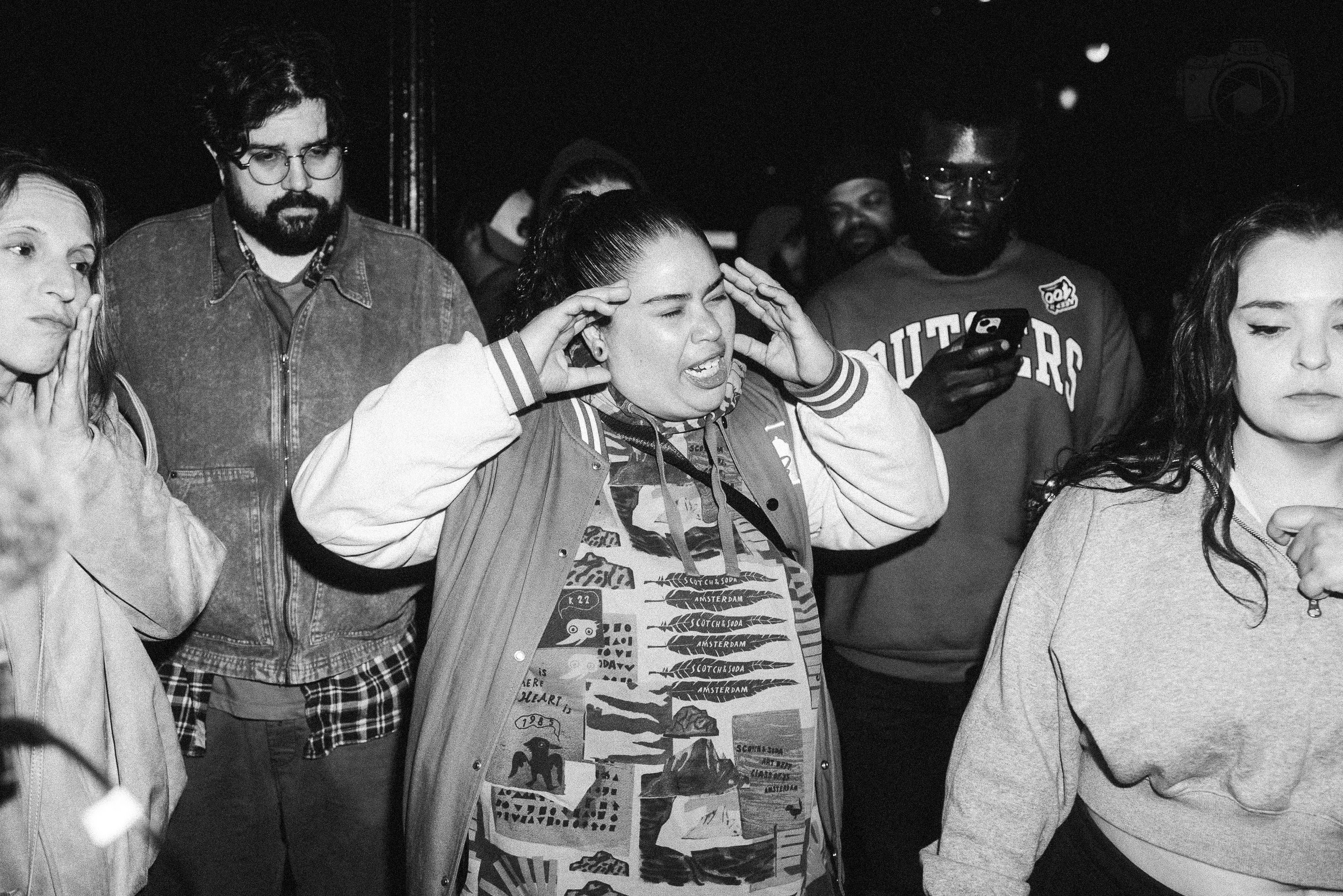 Group of people, mostly women, with one man, standing in a dark room, some with eyes closed or touching their heads, appearing to be engaged in a moment of concentration or emotion.