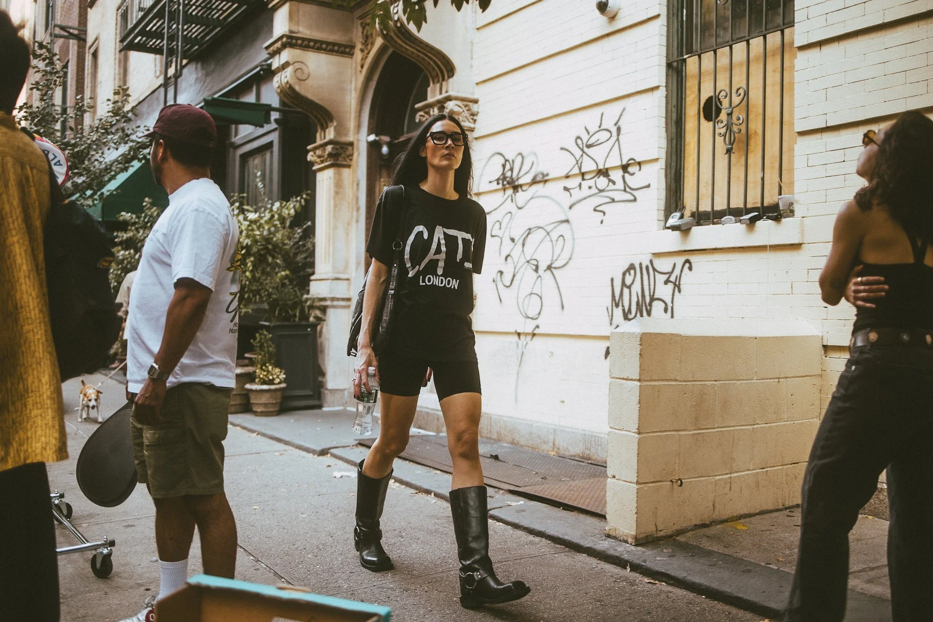 A woman with long dark hair wearing black glasses, a black t-shirt with words 'CAT LONDON', shorts, and black boots walking on a city street holding a water bottle. Several other people and a dog are nearby, and there is graffiti on the white brick w