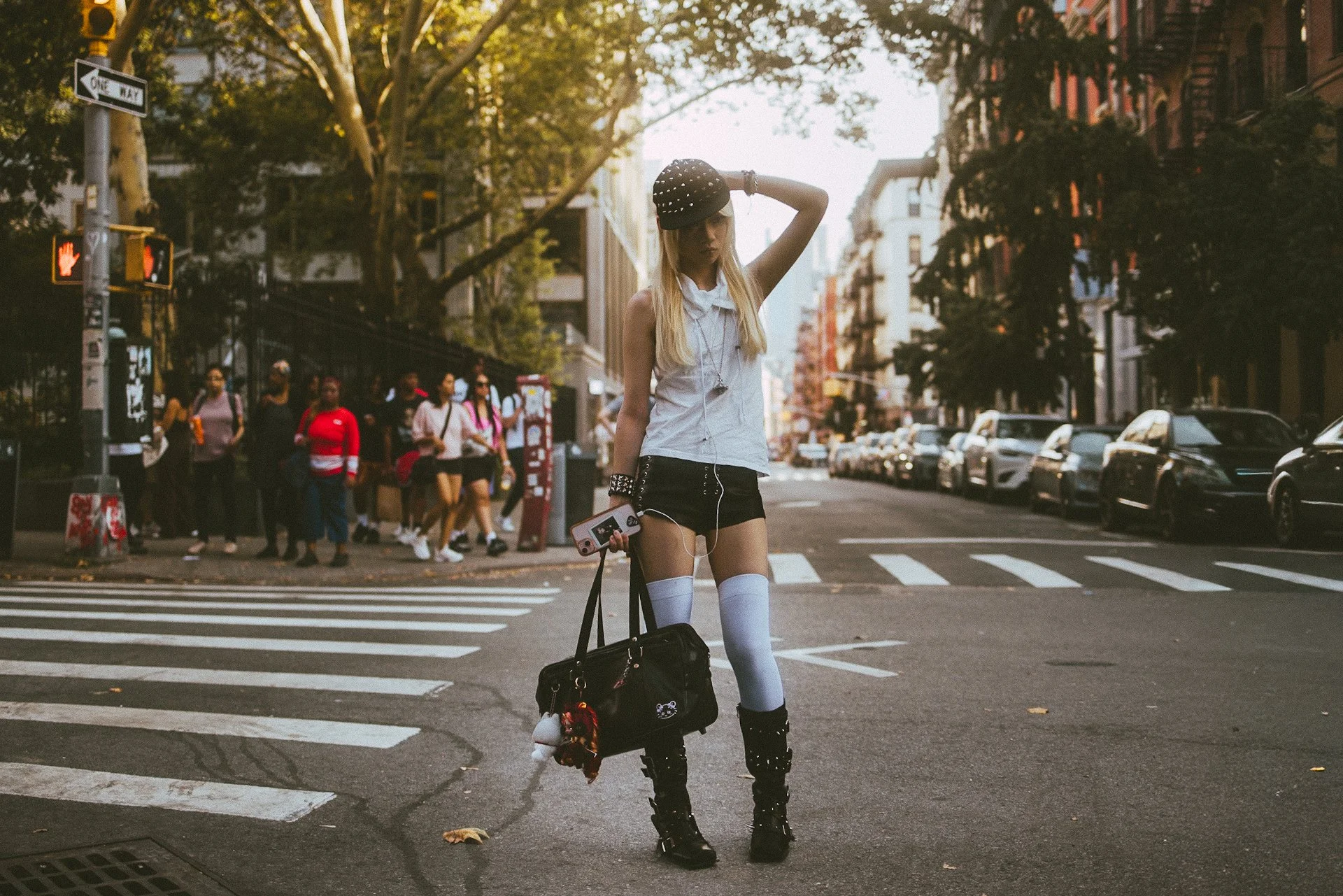 A young woman dressed in alternative fashion, wearing black boots, thigh-high socks, and a black hat, stands at a crosswalk in a city street while listening to music from her phone. She is holding a bag, and there are several pedestrians and parked c
